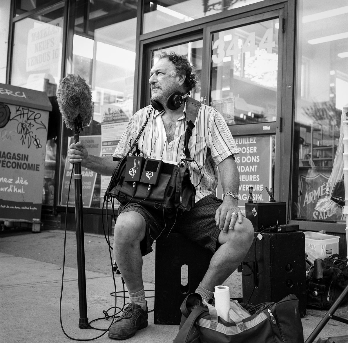 A man with headphones and audio equipment holds a microphone windscreen while sitting on the street outside a storefront.