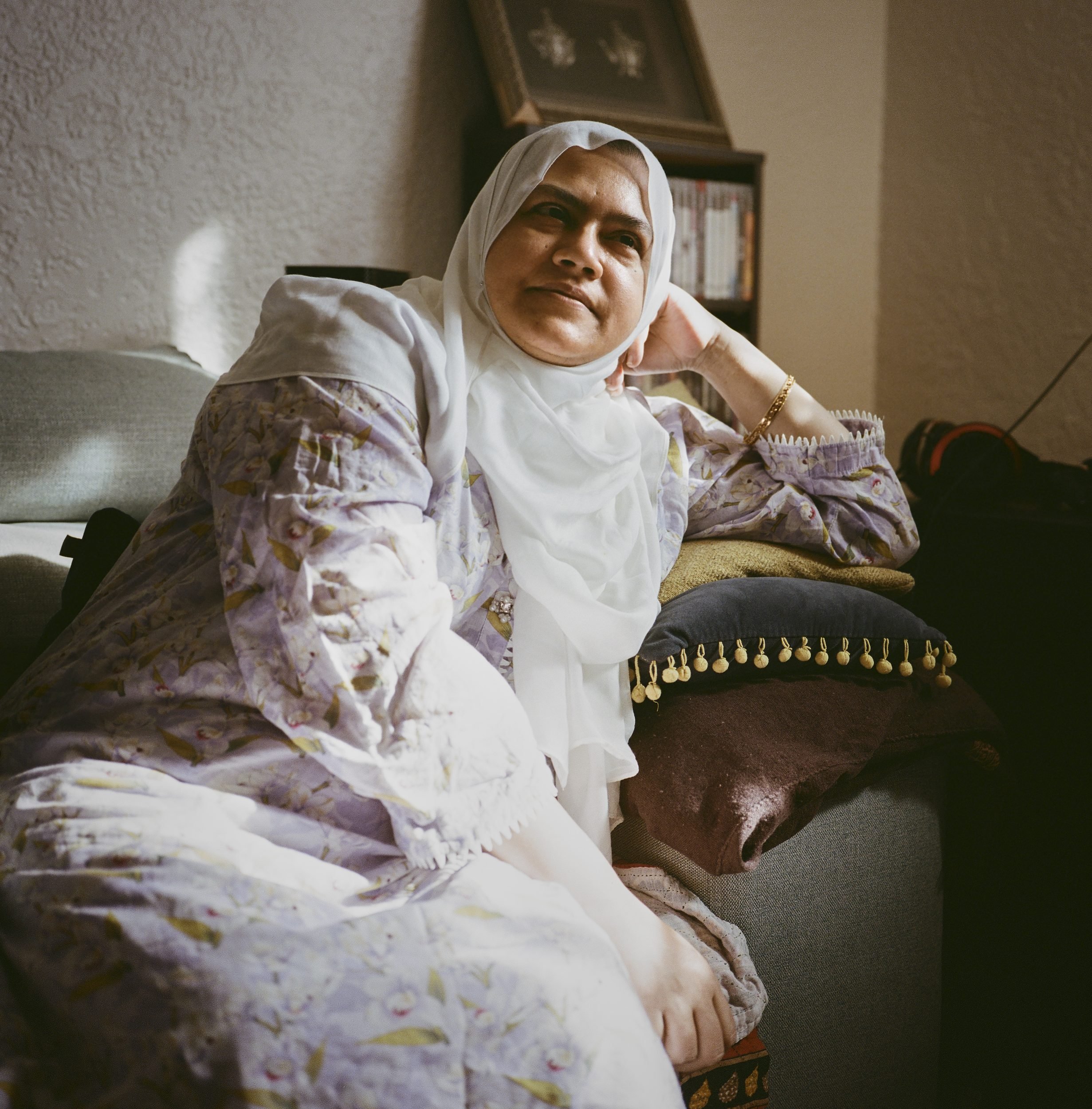 A woman wearing a white hijab and patterned jacket sits on a couch, resting her head on her hand.