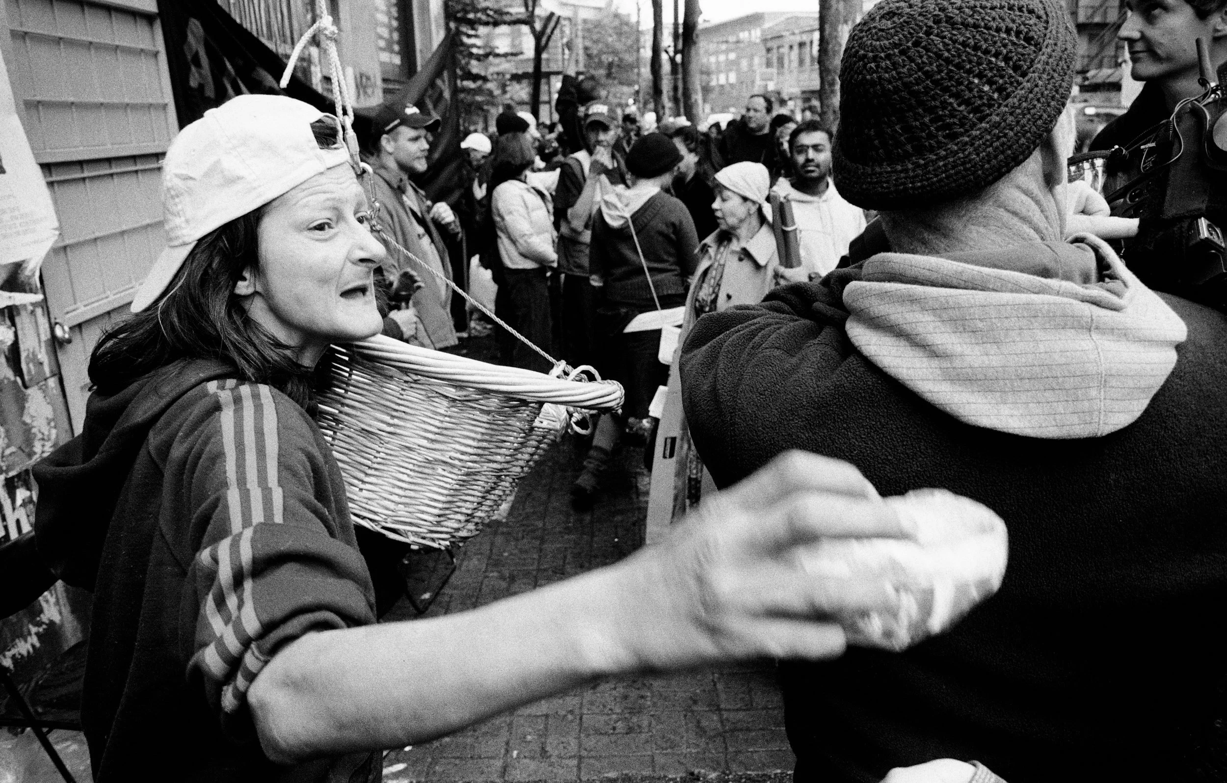 Woman in white cap holding basket speaks animatedly to man in crowd on urban street.