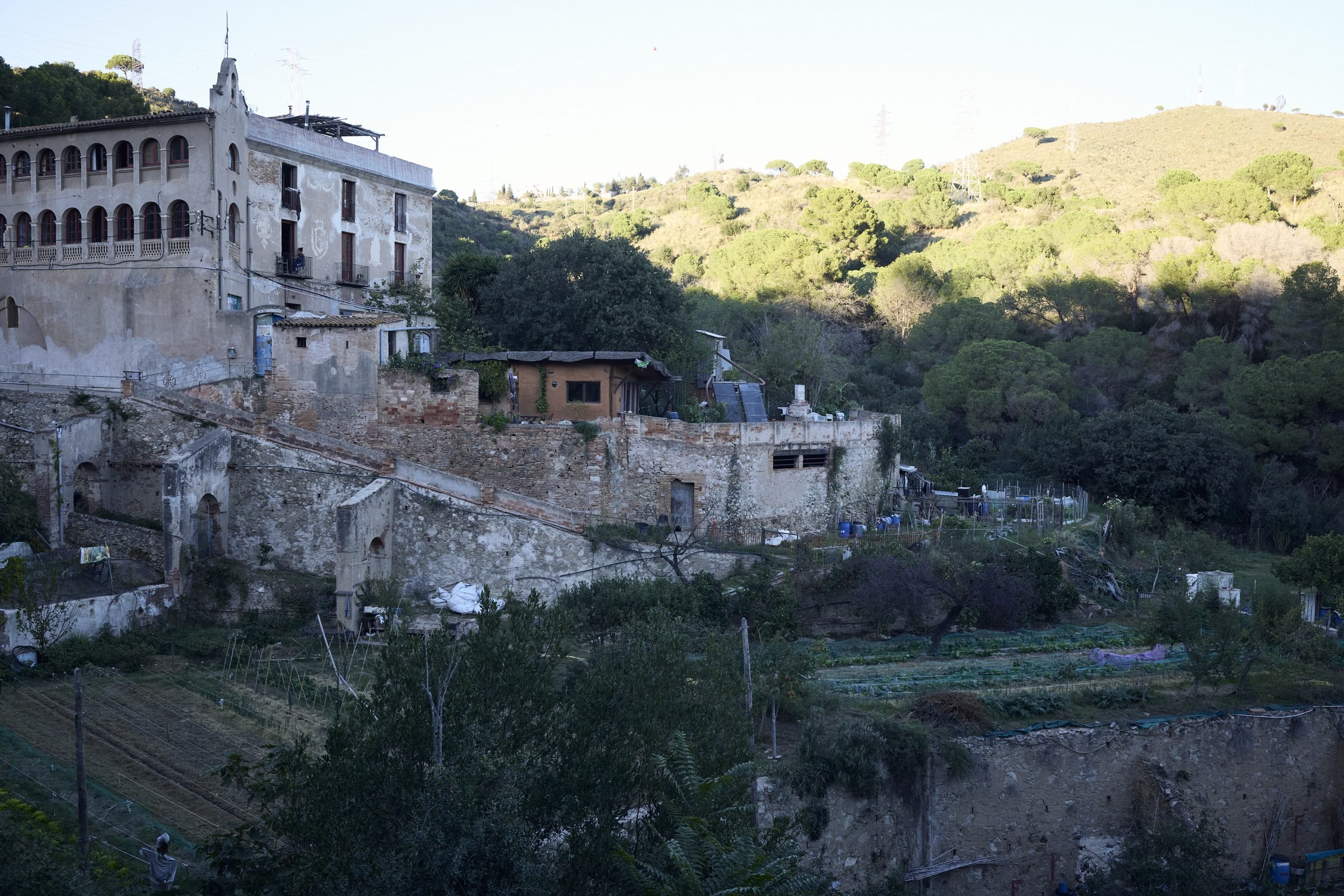 Hillside settlement with whitewashed colonial-style buildings, vegetable gardens, and forested slopes.