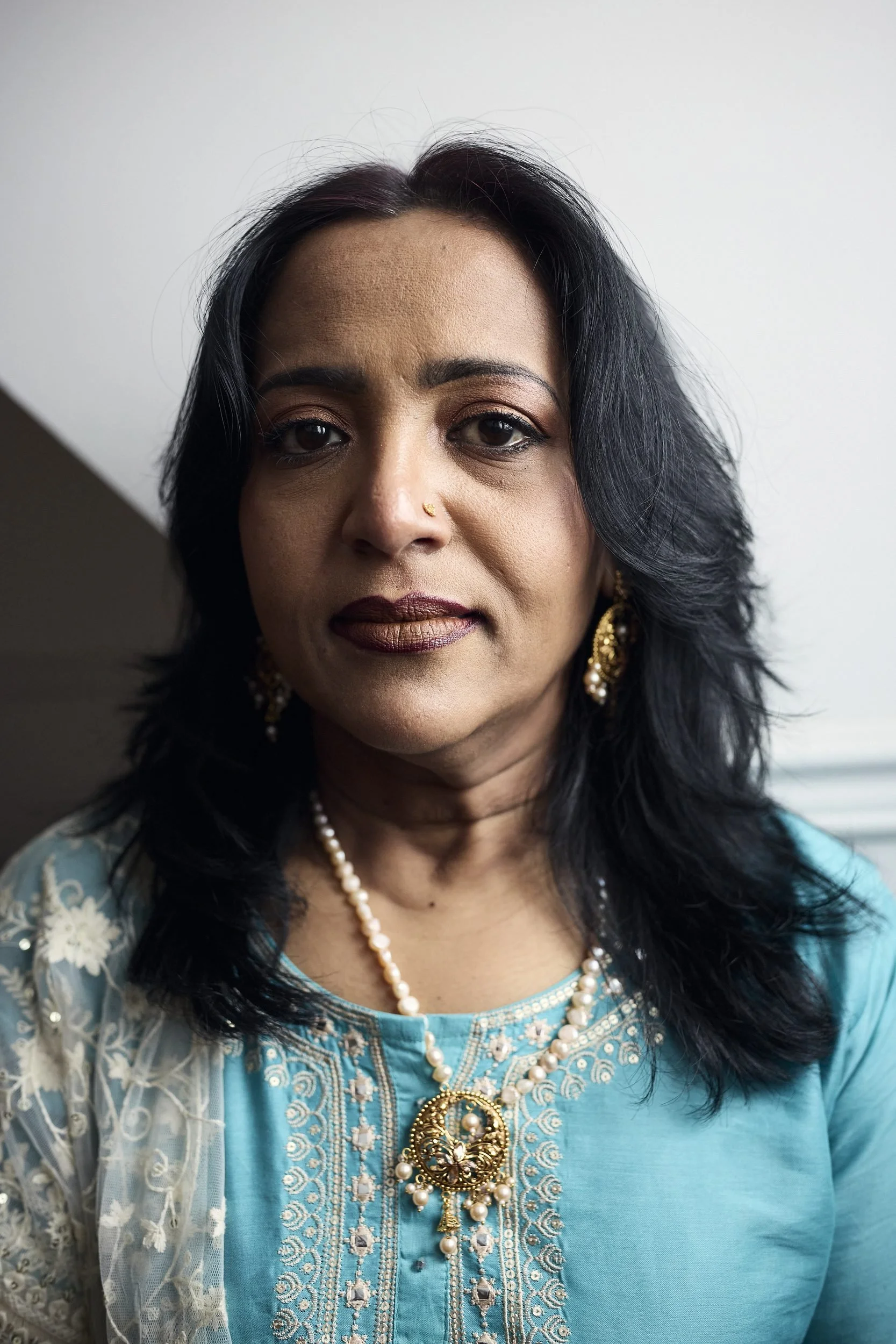 Woman in turquoise embroidered tunic and pearl jewelry looks toward camera in studio setting.