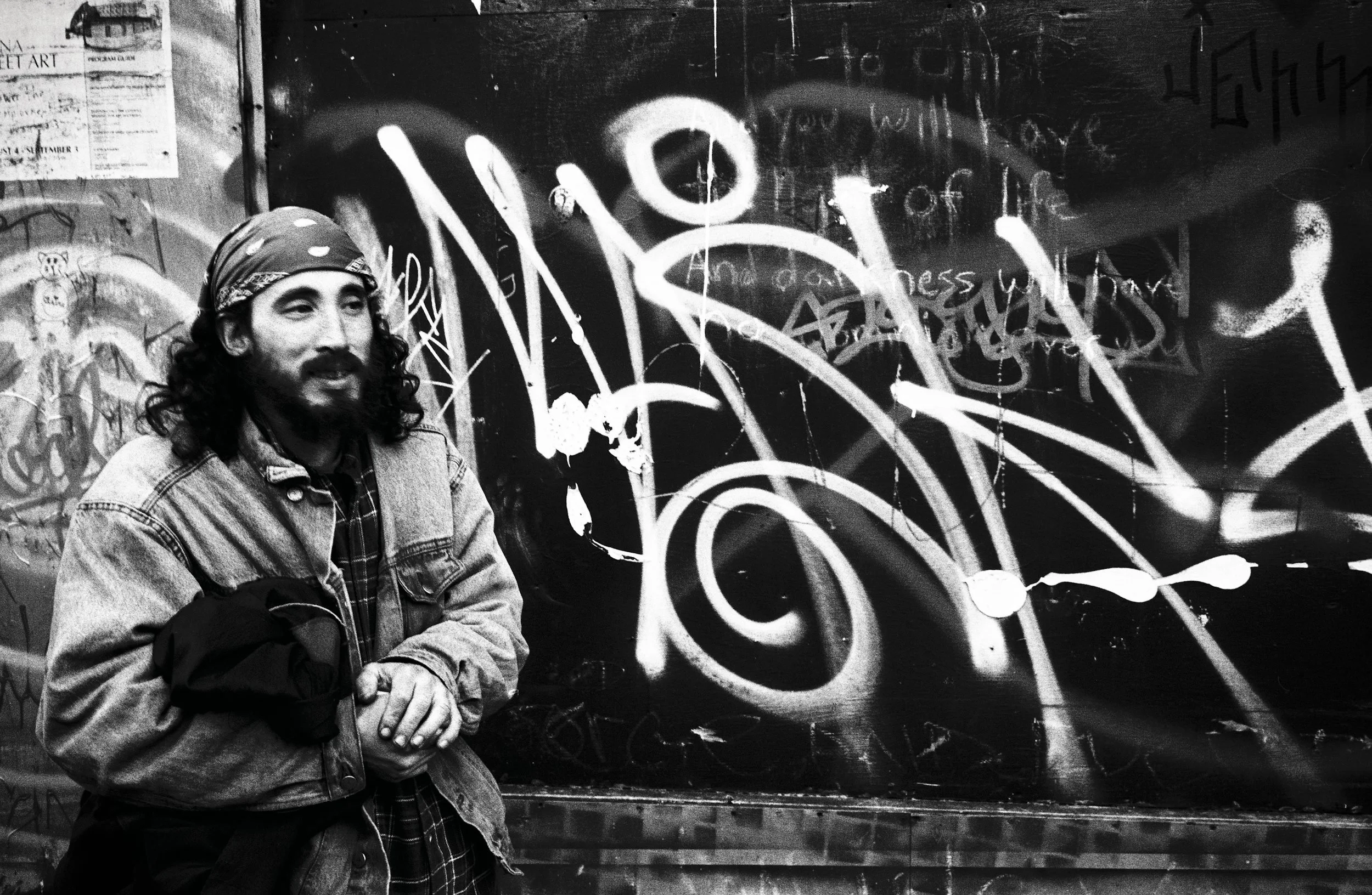 Man with headband and beard stands before a wall covered with white graffiti and text.