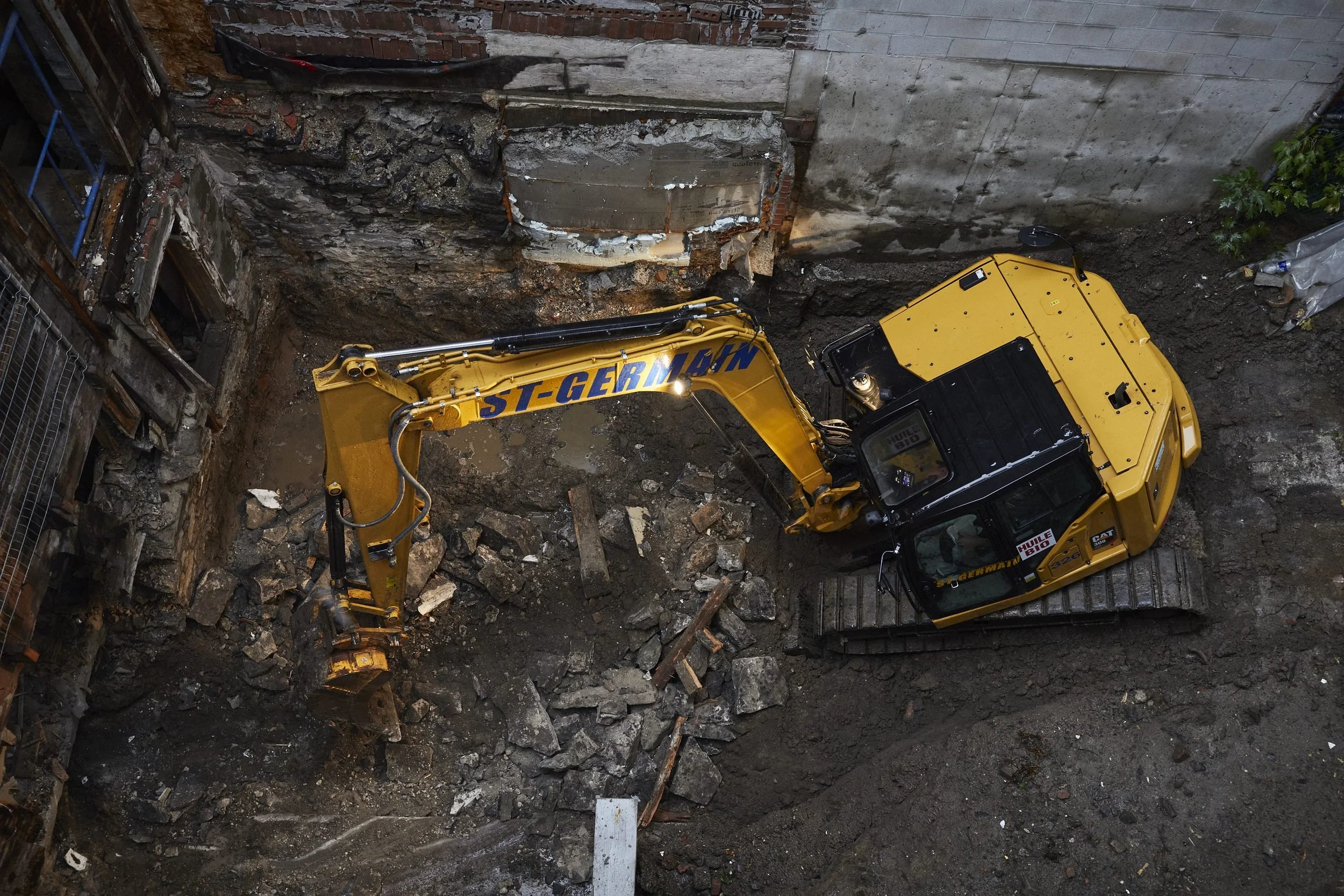 Aerial view of a yellow excavator and dump truck working in a confined urban alley with deteriorated buildings.