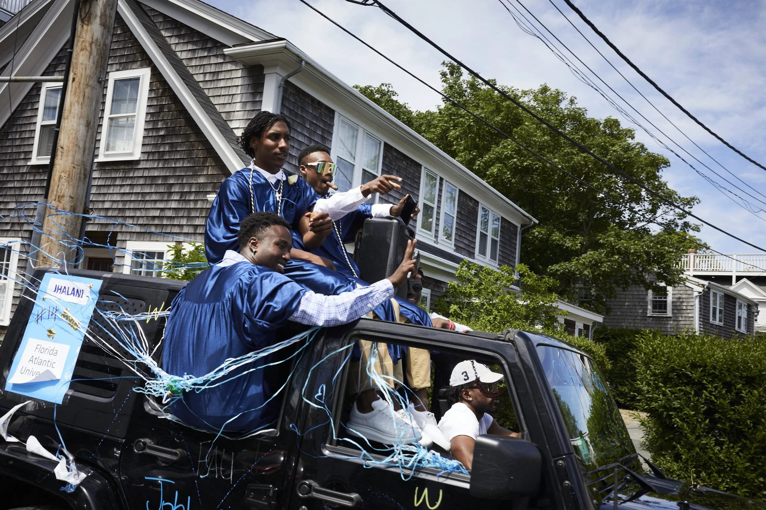 People in blue jackets wave from a decorated vehicle during what appears to be a parade in a residential neighborhood.