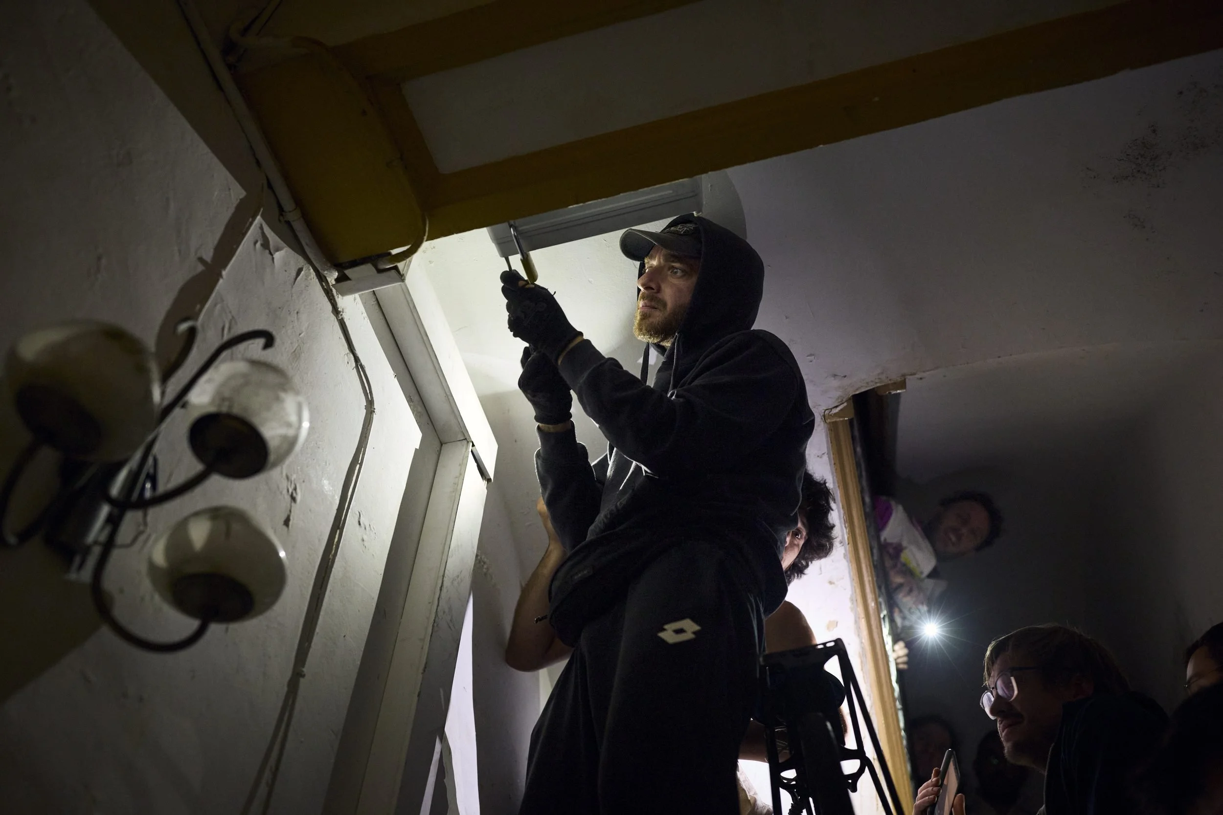 A man in dark clothing and cap works on ceiling fixtures while others watch from below in an indoor space.