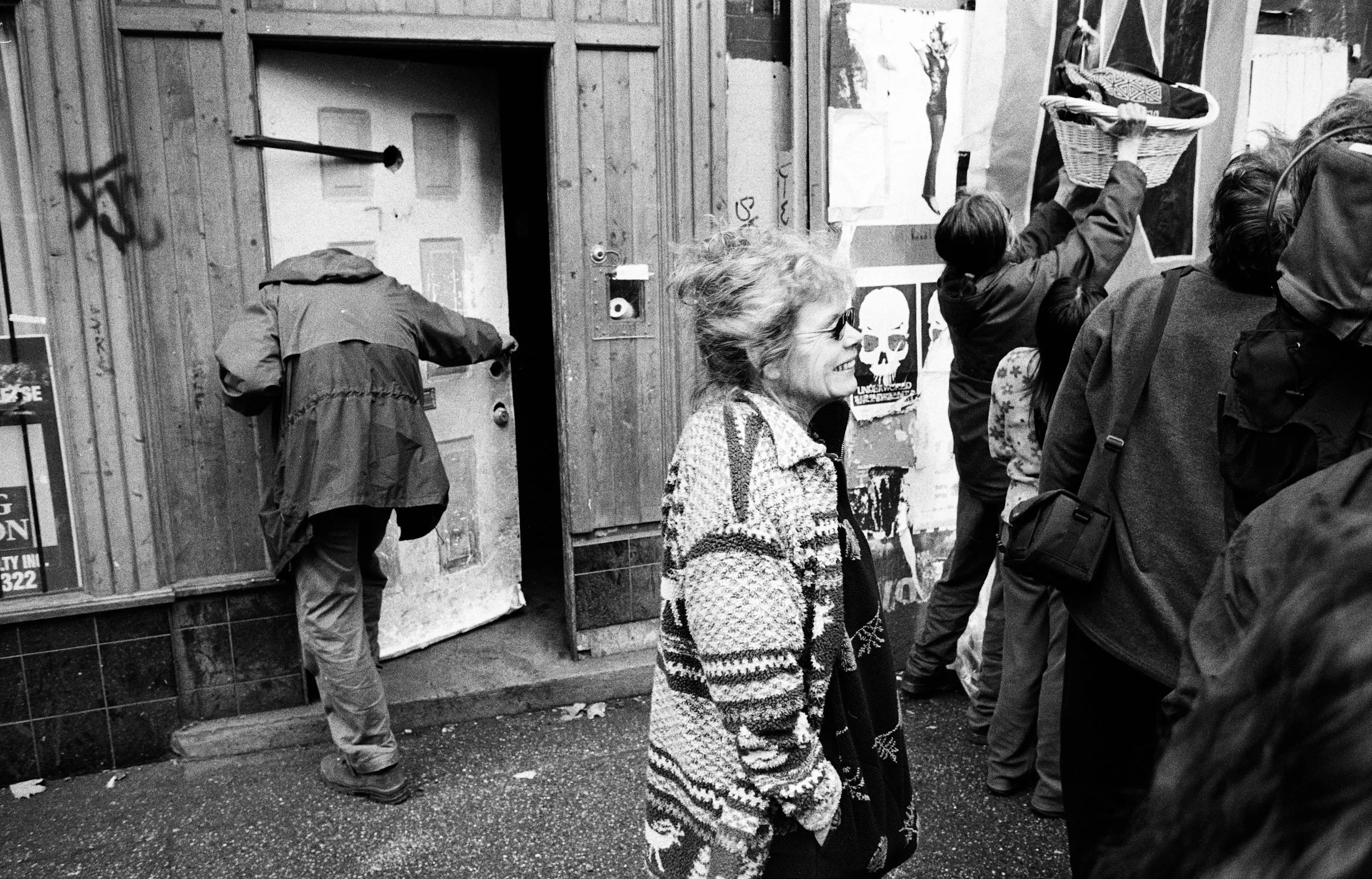 A woman in a patterned cardigan stands among a group of people gathered outside a weathered building entrance.