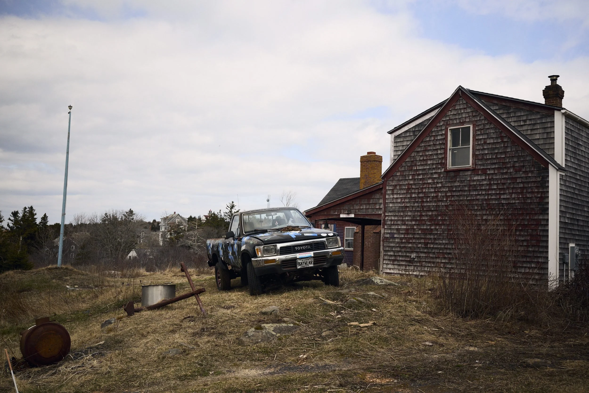 A weathered Toyota pickup truck parked beside a shingled cottage on an overgrown rural property.
