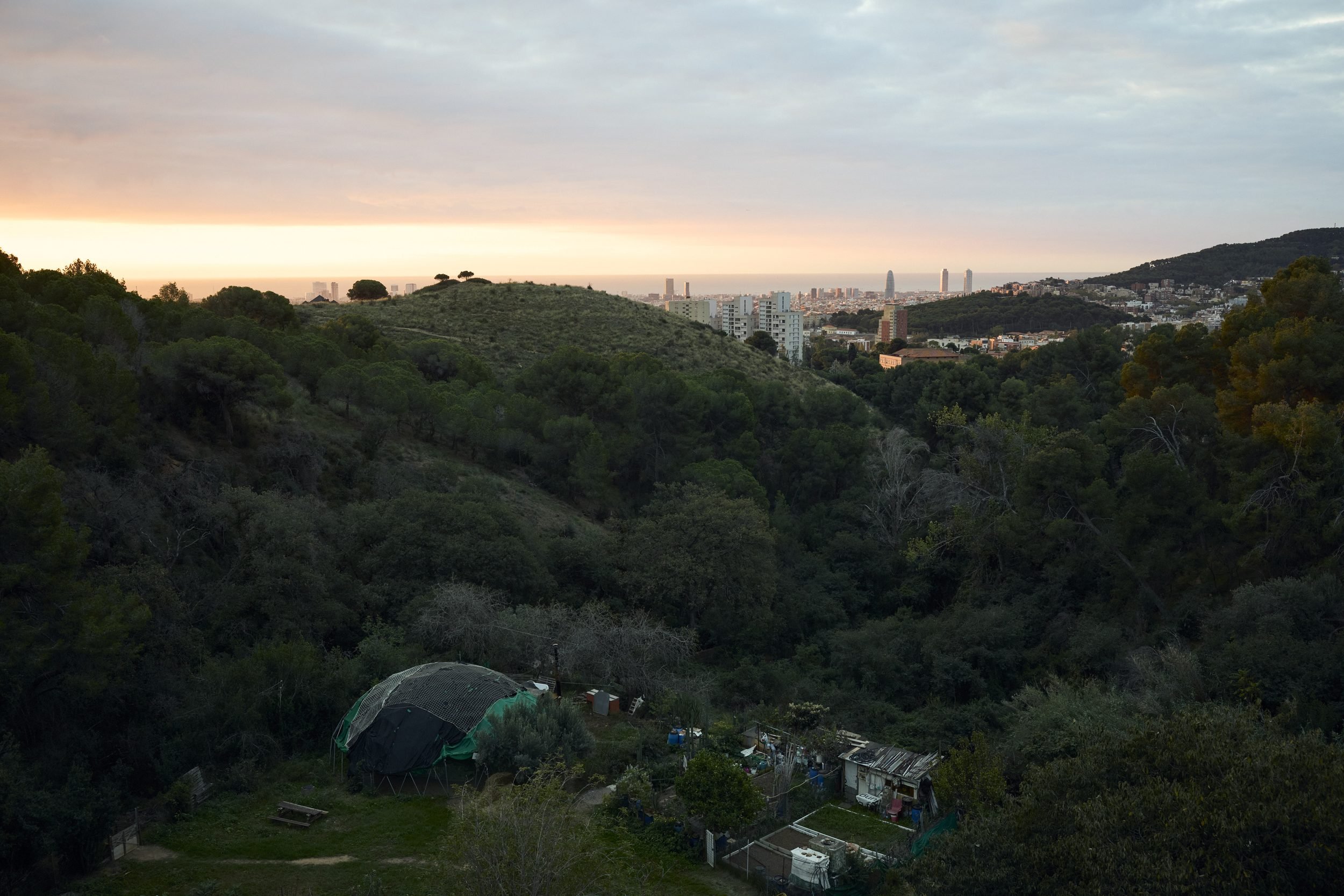 A large geodesic dome structure sits in a forested valley with a city skyline visible in the distant background at dusk.