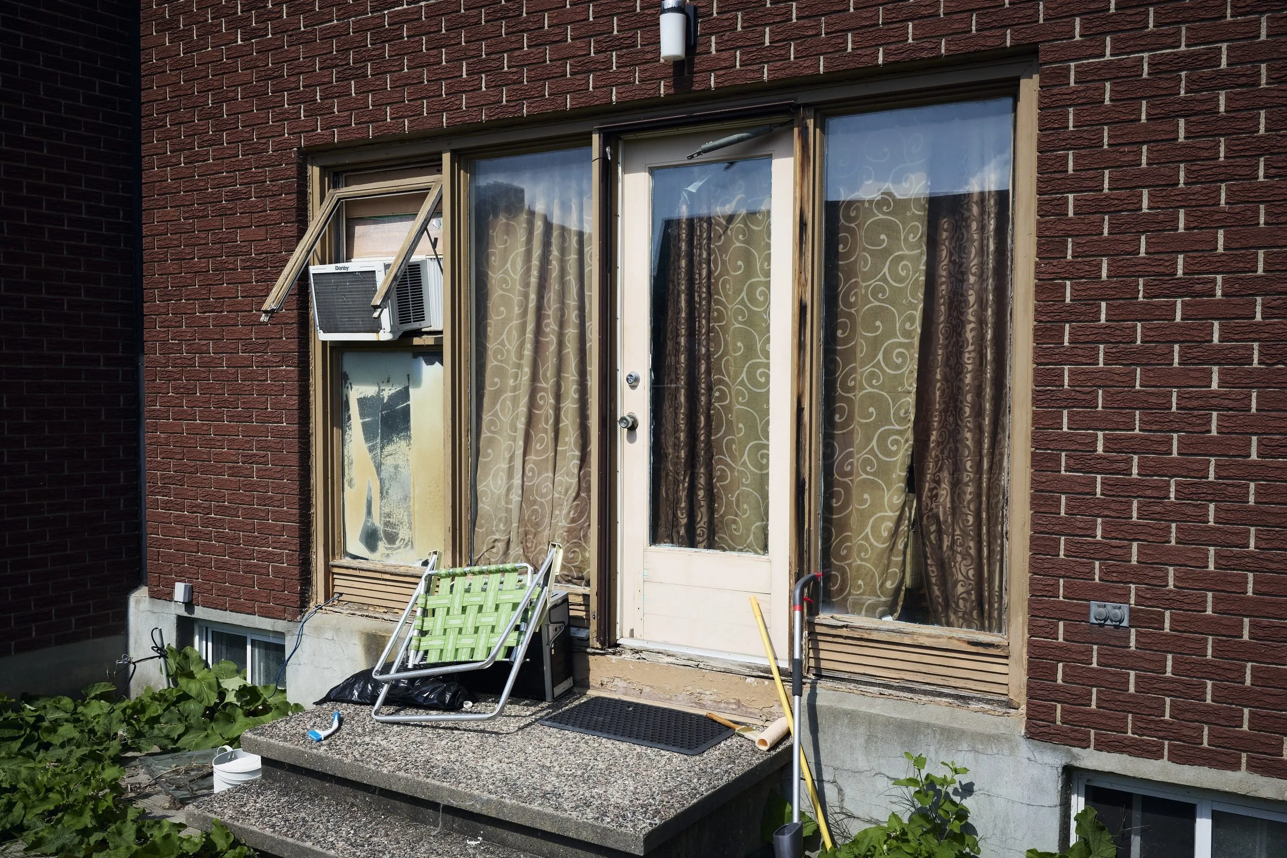 Brick residential building entrance with glass doors, patterned curtains, window air conditioner, and lounge chair on concrete patio.