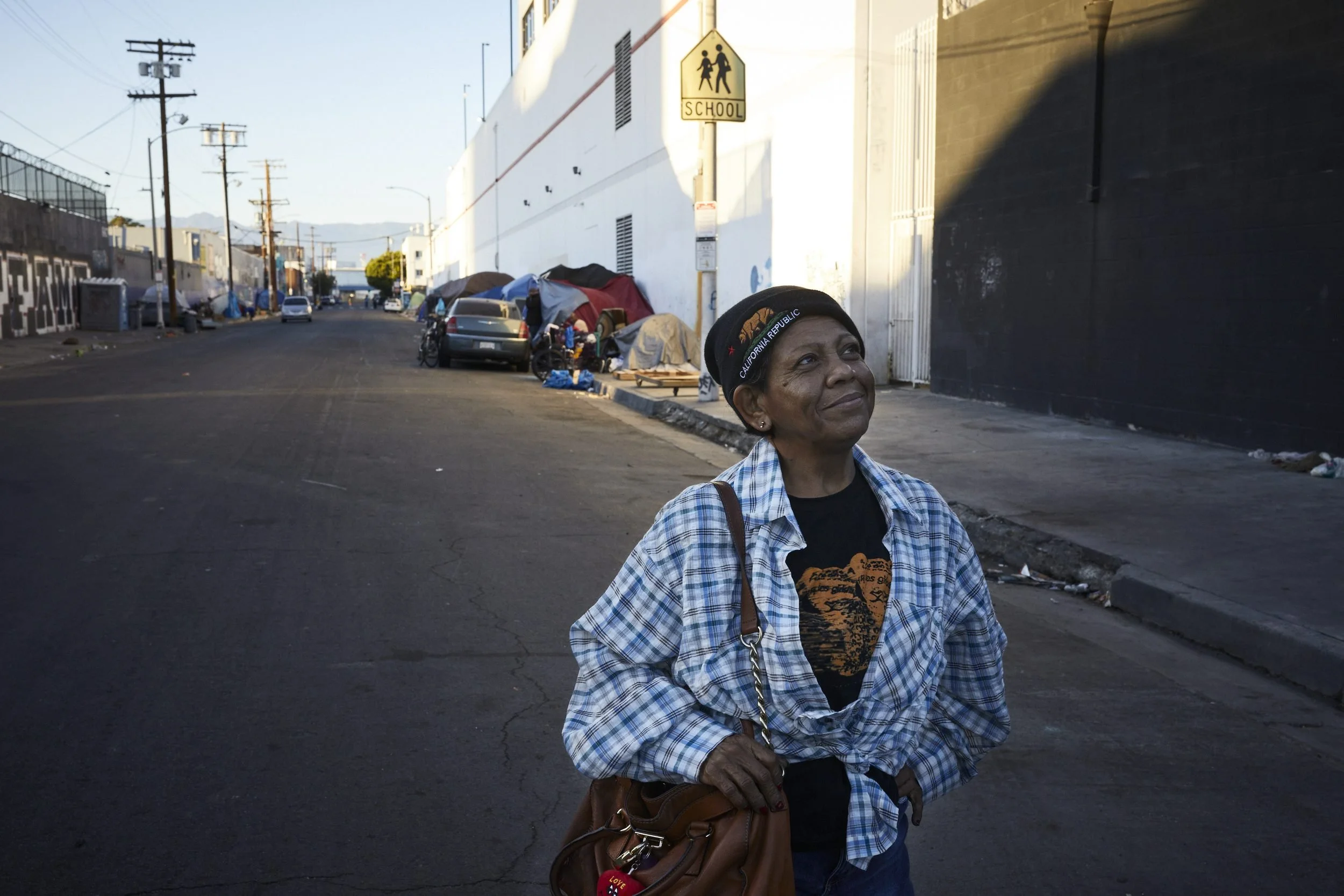 A woman in a blue plaid shirt stands on a street near homeless encampments and industrial buildings.