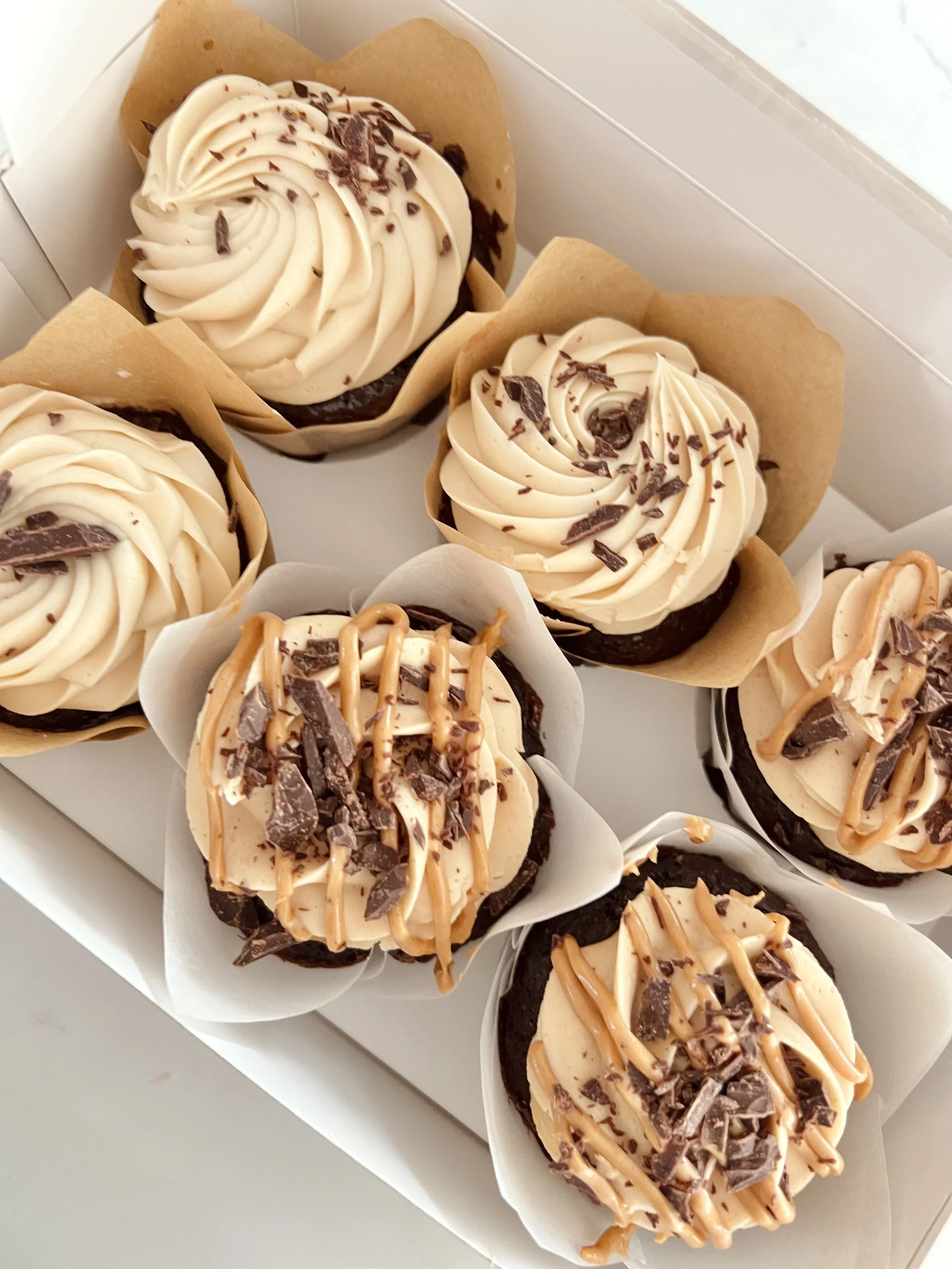 Close-up of vanilla cupcakes topped with chocolate shavings, drizzle, and piped frosting, wrapped in bakery paper holders.