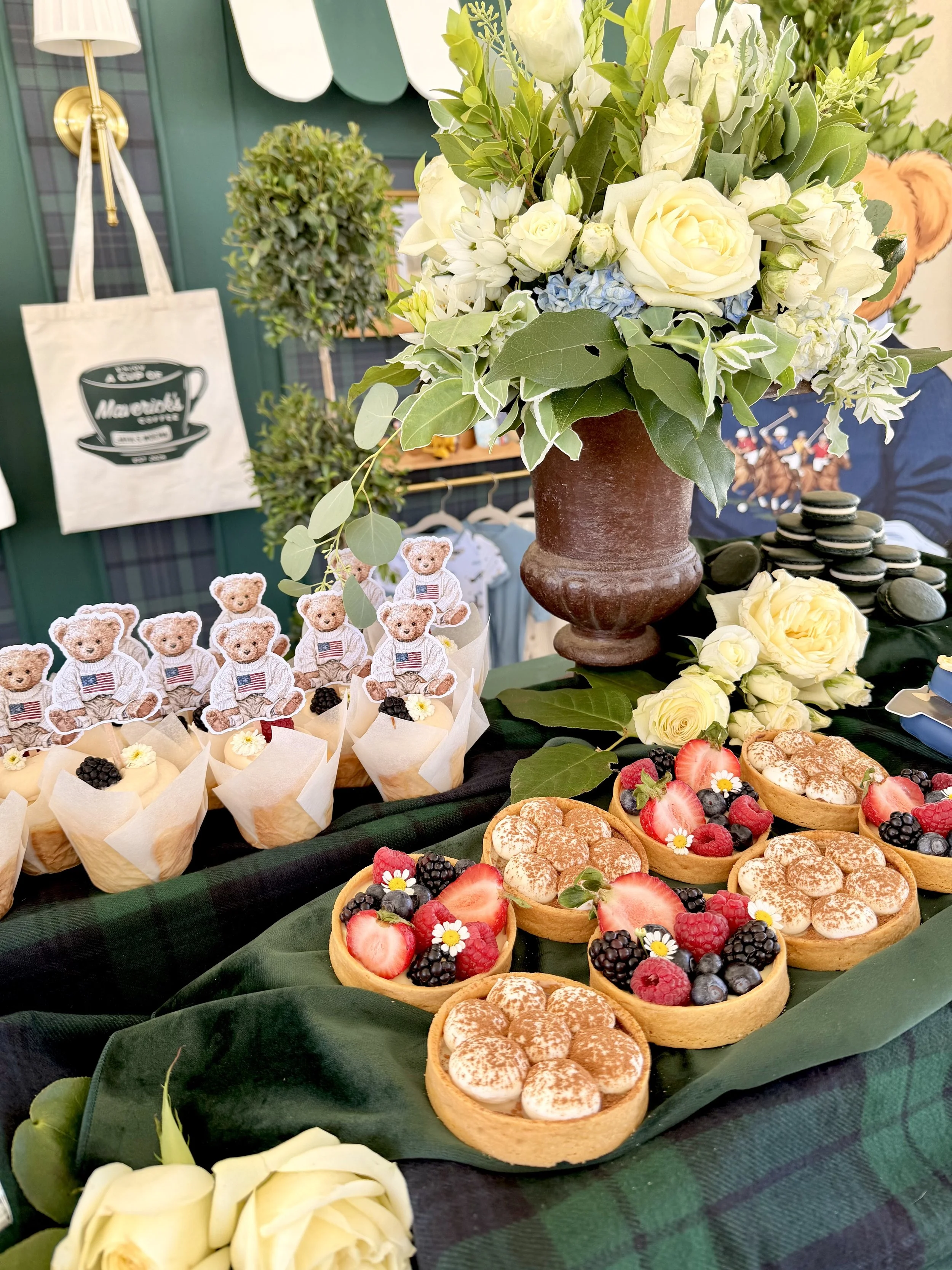 A display of assorted fruit tarts topped with strawberries, blackberries, raspberries, and edible flowers. A large floral arrangement with white roses, green leaves, and other white flowers stands behind the tarts. In the background, there are decorative items, including a tote bag with a coffee cup design and a small topiary plant, on a dark green tablecloth.