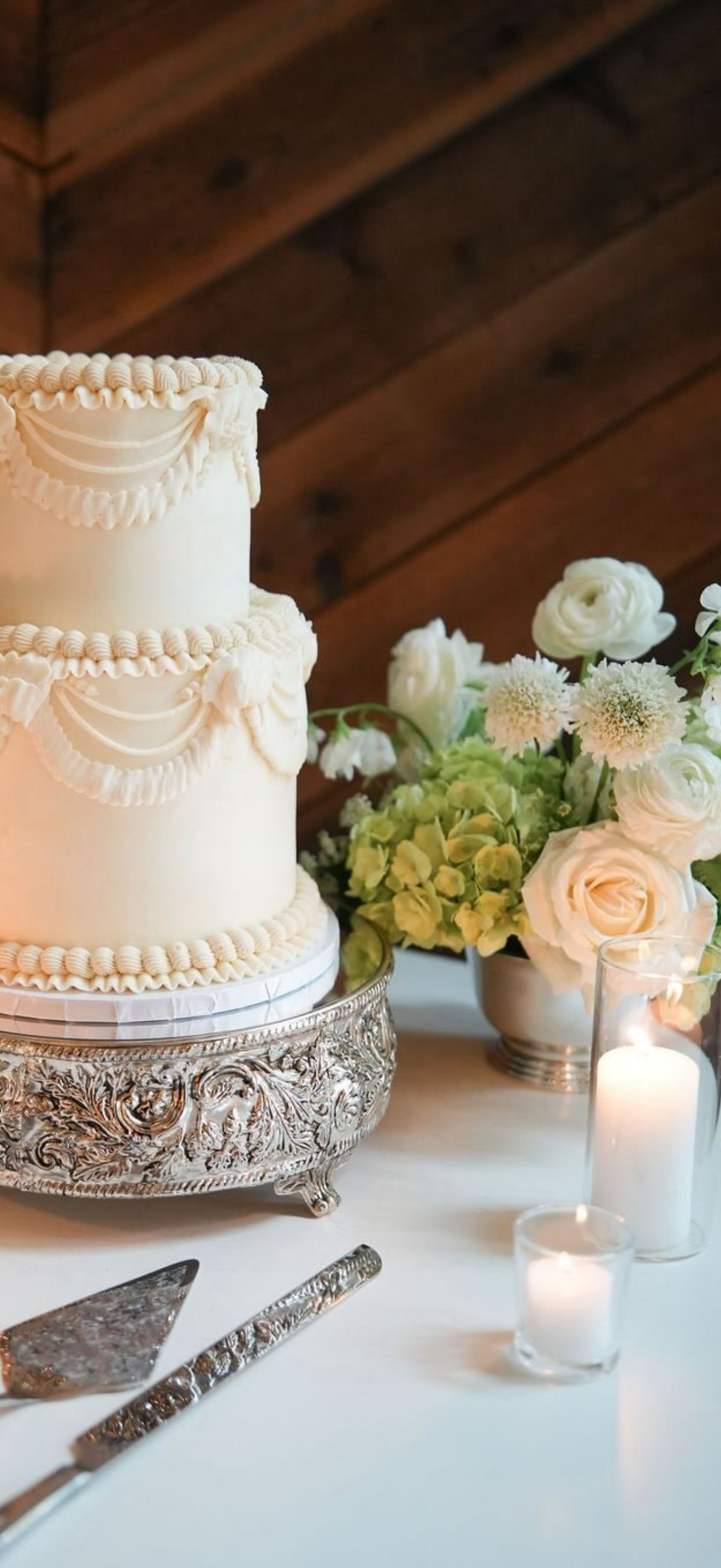 A white, three-tiered wedding cake with decorative piping and draping sits on an ornate silver cake stand. Next to the cake, there are white and pale yellow flowers in a white vase, and candles in glass holders emit a soft glow.