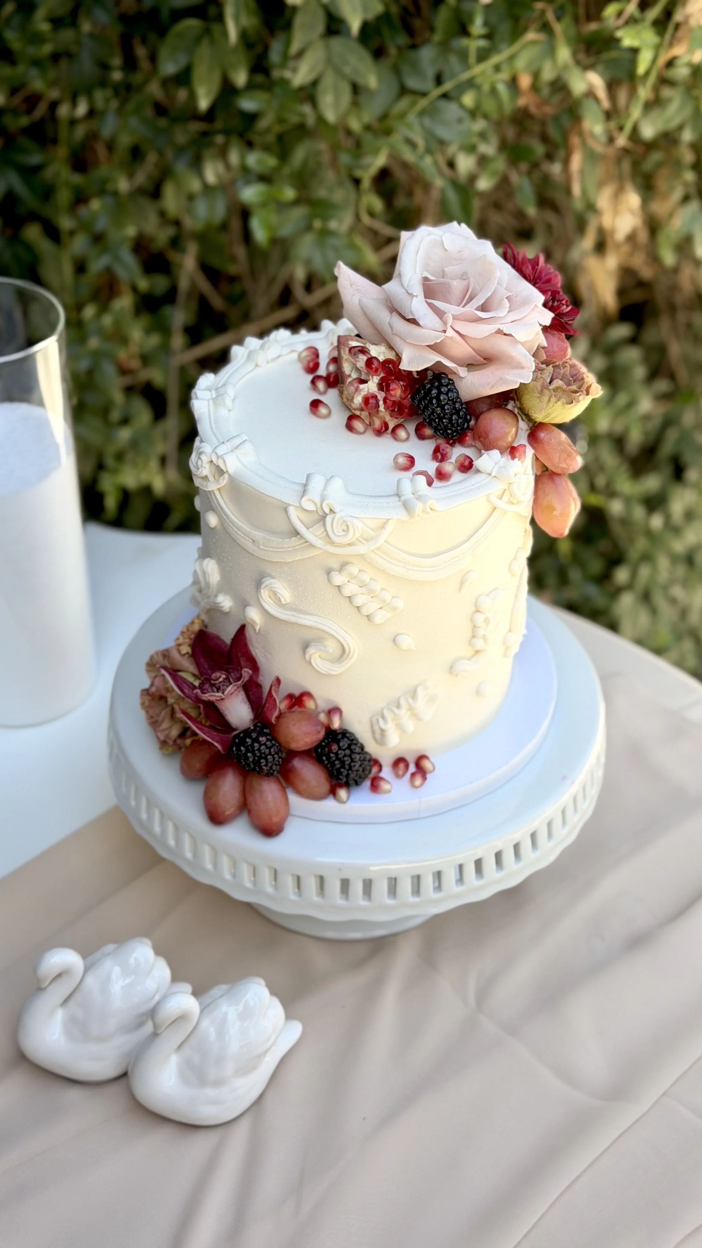 Elegant white wedding cake decorated with fresh flowers and berries, placed on a white stand, with a glass of milk nearby, outdoors with greenery in the background.