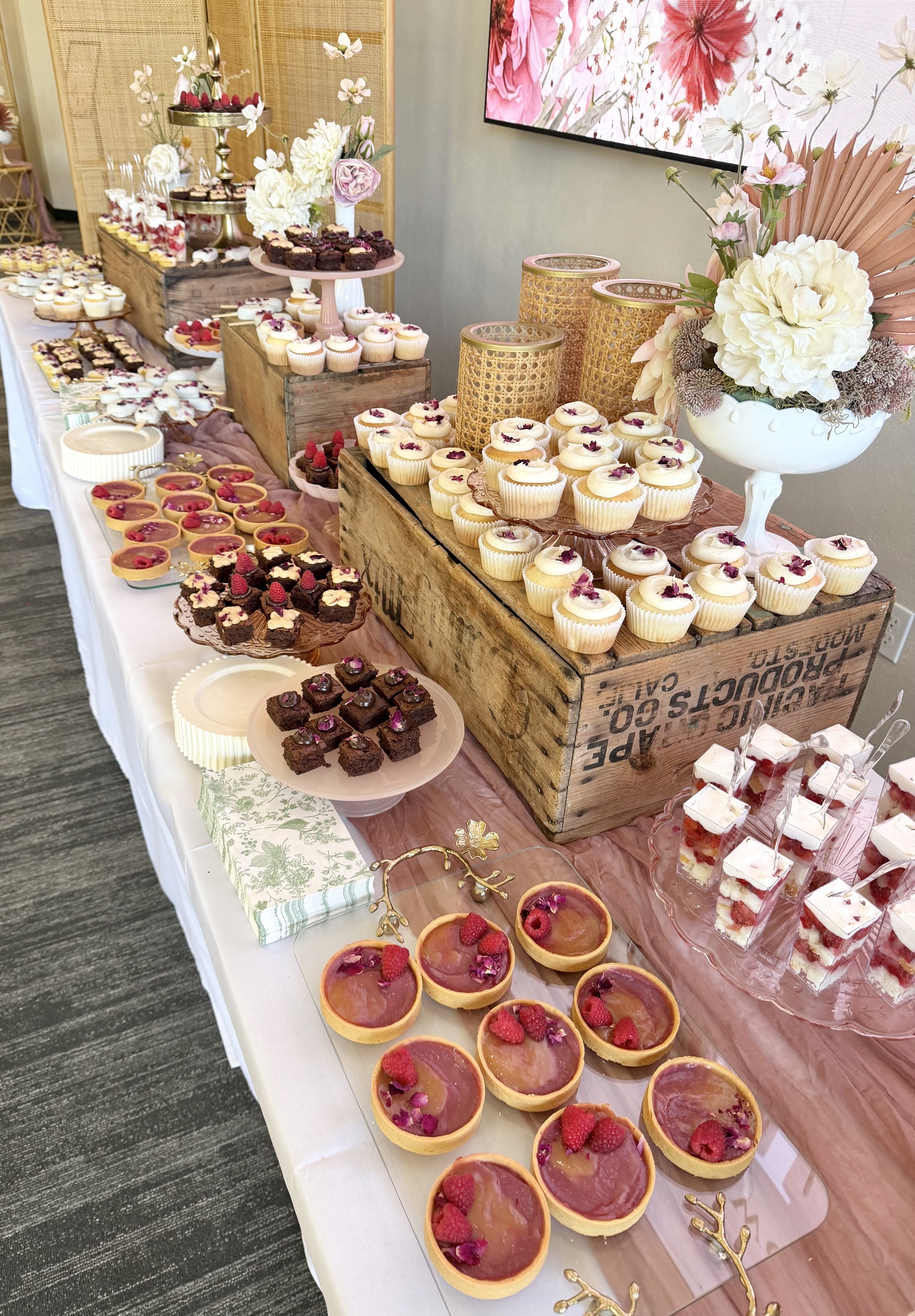 A dessert buffet table with an assortment of cupcakes, tarts, and parfaits, decorated with flowers and gold accents, displayed on wooden crates and trays.