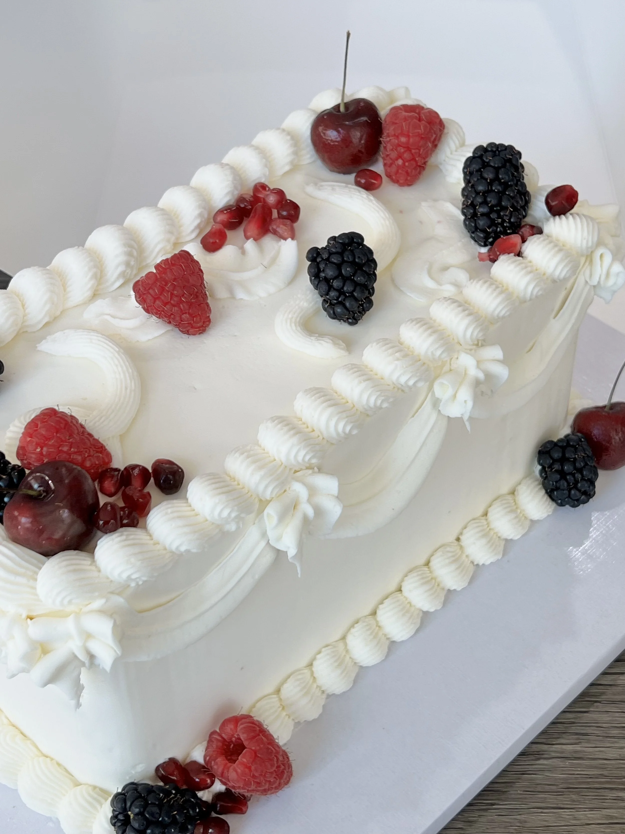 Close-up of a white frosted rectangular cake decorated with fresh berries including cherries, raspberries, blackberries, and pomegranate seeds, with piped white frosting edges.