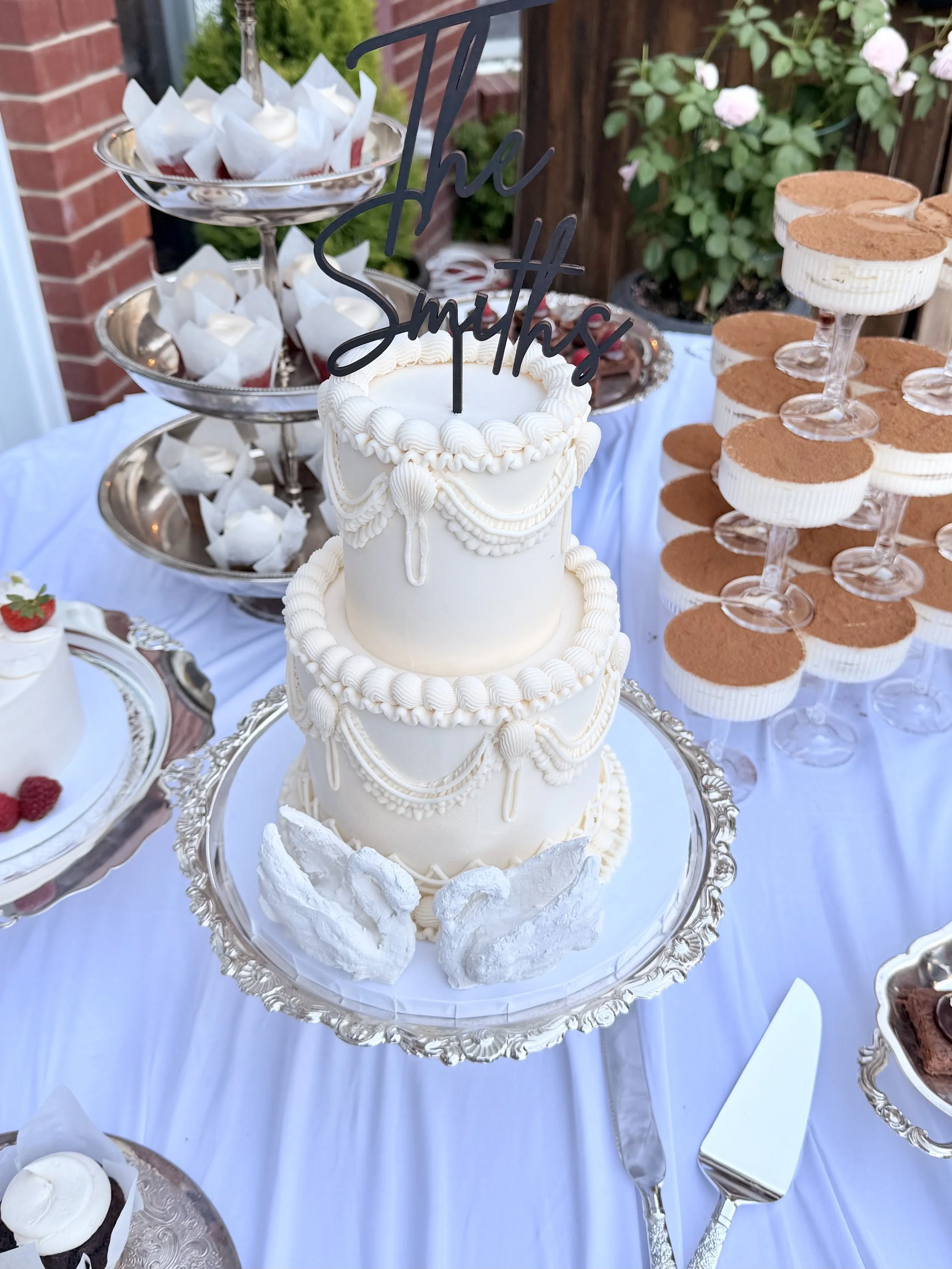 Three-layer white wedding cake decorated with piped frosting and seashell designs, topped with a black "And They Said" cake topper, placed on a silver cake stand surrounded by meringue cookies and other desserts, with a garden background.