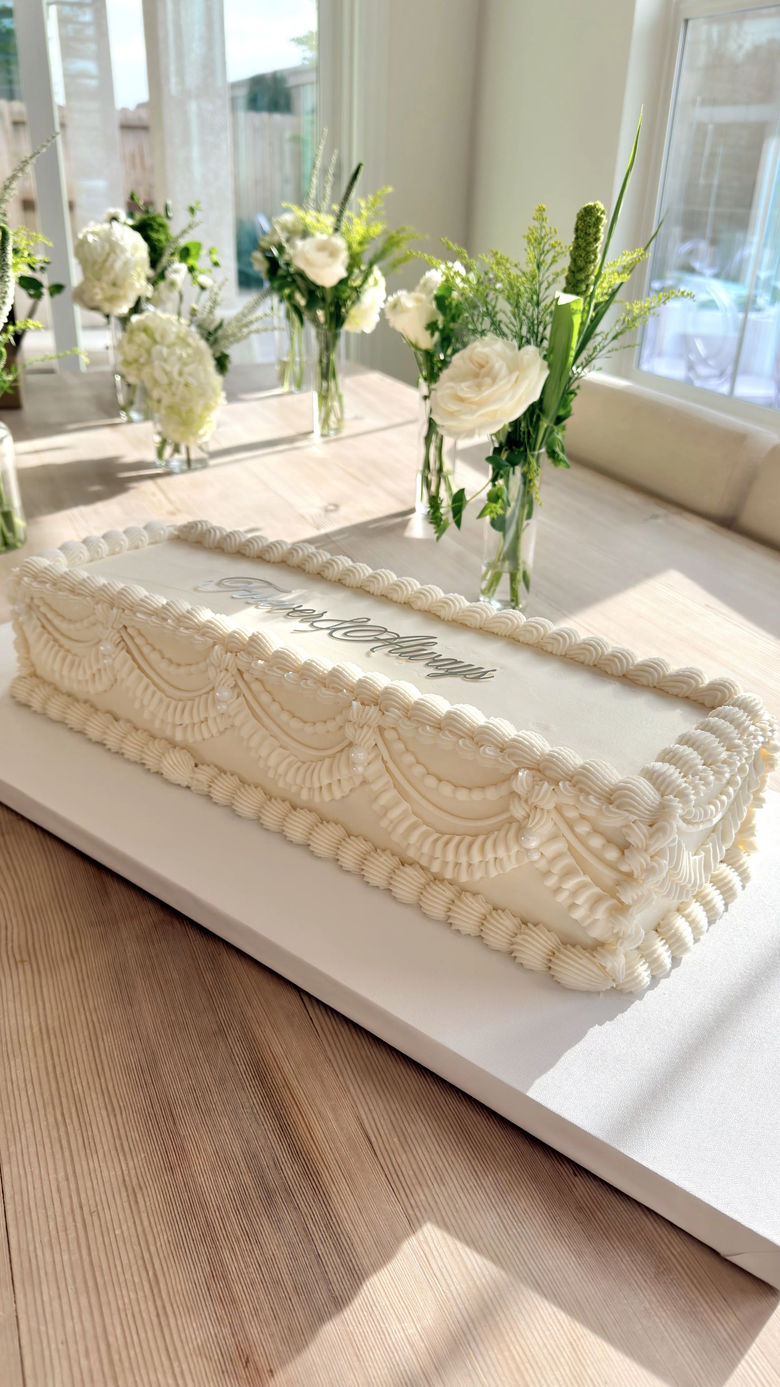 A rectangular white cake with decorative piping and pearls, with the word 'Congratulations' written on top, placed on a white board on a wooden table. In the background, there are several glass vases with white flowers on a table near a window.