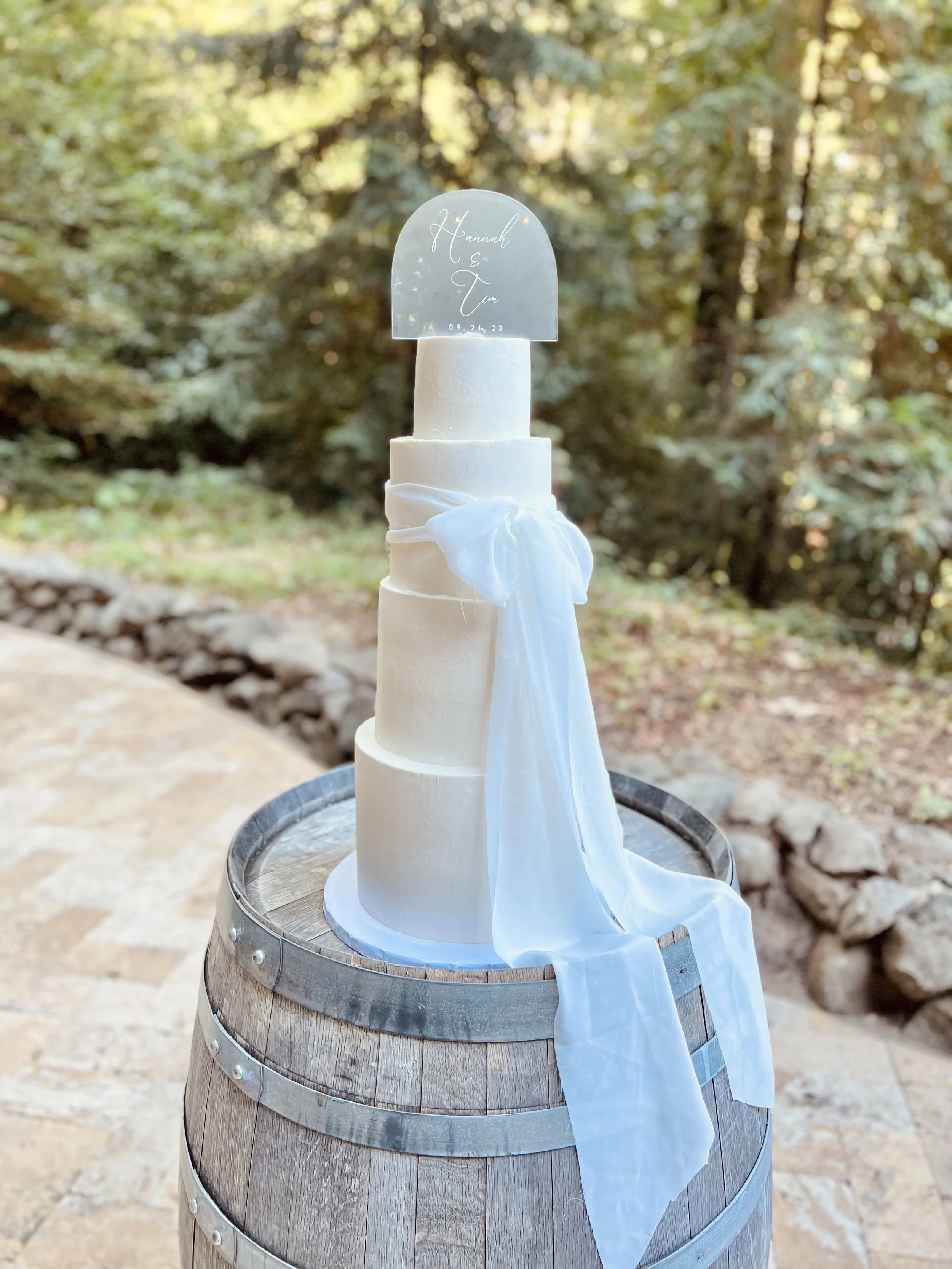 A three-tiered white wedding cake decorated with white fabric, placed on a wooden barrel outdoors. A transparent acrylic topper with handwritten-style text reading 'Hannah & Tim 09.24.23' is on top.