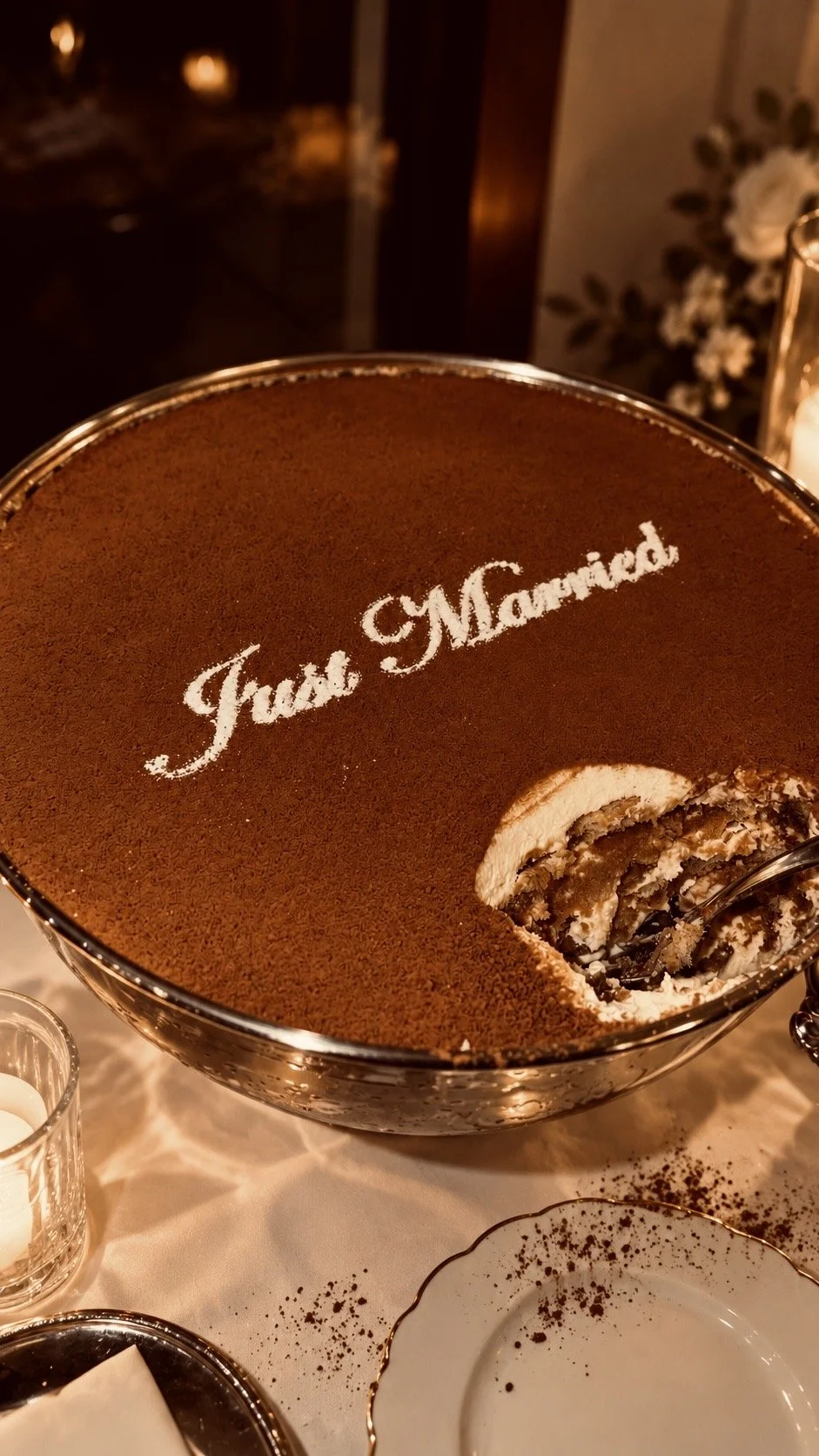 A large bowl of tiramisu dessert with the words "Just Married" written on top in powdered sugar. A spoon is scooping some of the tiramisu.