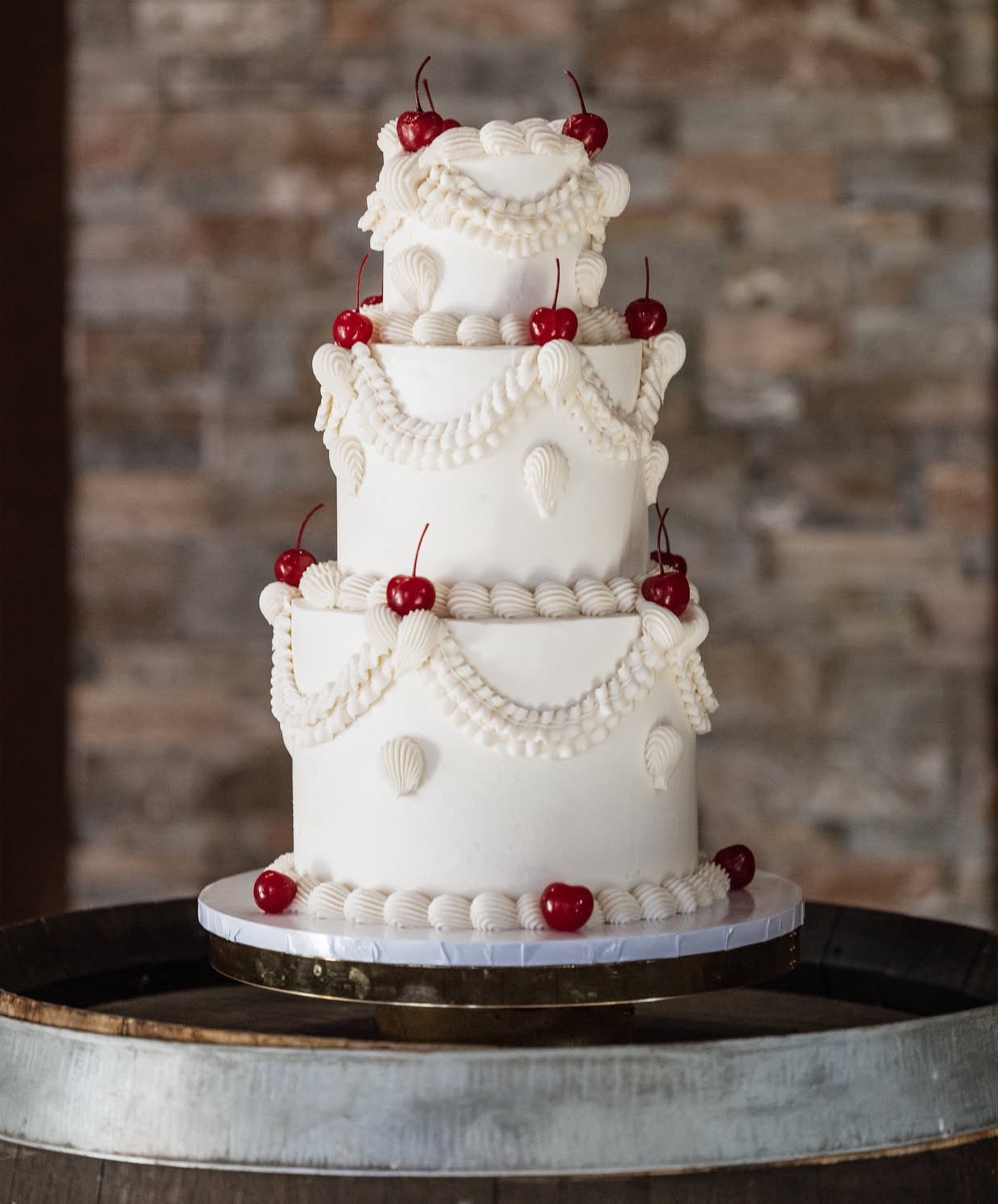 A four-tier white wedding cake decorated with white icing swags and shells, topped with cherries, on a silver cake board.