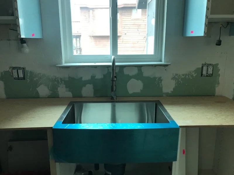 New kitchen sink installed beneath a window, with unfinished drywall walls and electrical outlets.