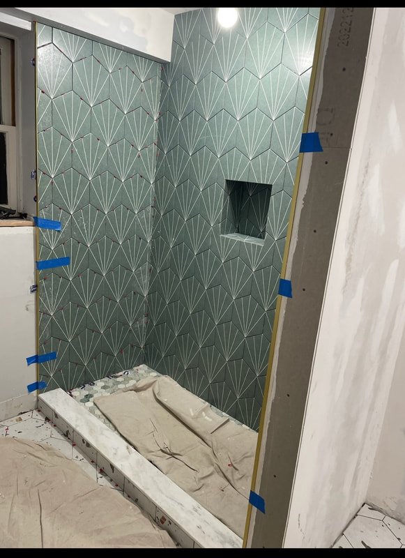 A bathroom shower area under construction with geometric patterned blue-green tiles on the wall, a built-in niche, and protective coverings on the floor and edges.