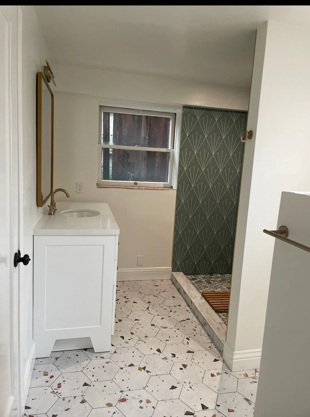 Bathroom with a white vanity, a mirror, a window, and a patterned shower area with a pebble floor and wooden bath mat.