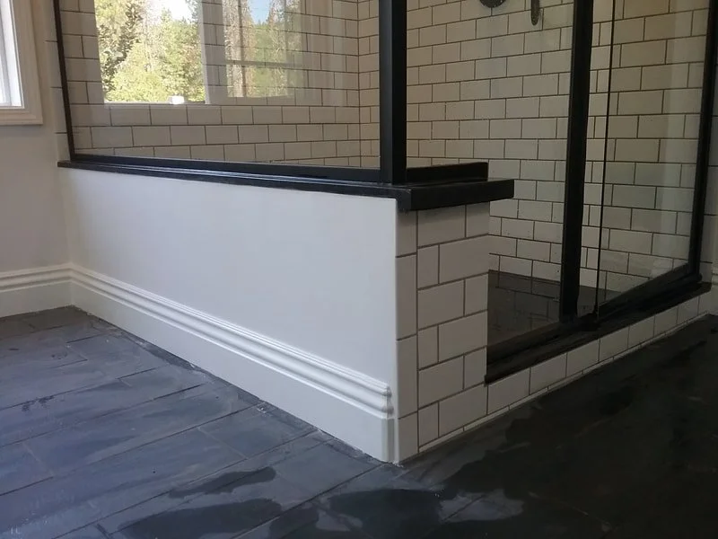 Interior corner with a white half wall, black trim, and brick-patterned tiles surrounding an enclosed shower area. The floor has dark tiles and a window showing trees outside.