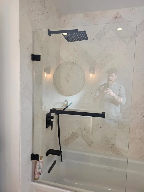 Bathroom shower with a glass door, modern black fixtures, a rainfall showerhead, a handheld shower, and a round mirror on marble wall, with a person taking a photo reflected in the glass.
