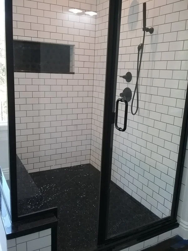 Walk-in shower with black fixtures, white subway tile walls, black hexagonal tile floor, and a built-in niche for toiletries, all enclosed with a glass door.