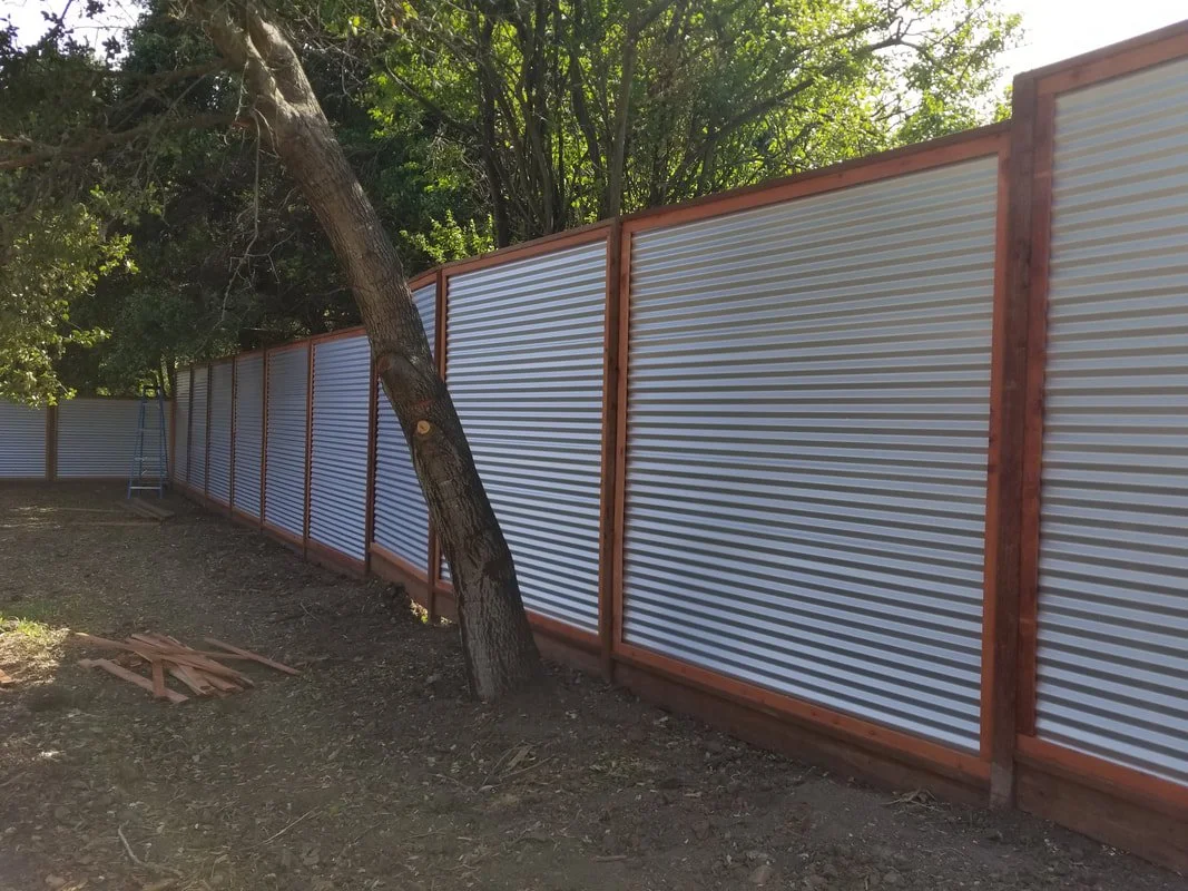A long metal fence with a wooden frame, situated next to a leaning tree in an outdoor area with dirt ground and green trees in the background.