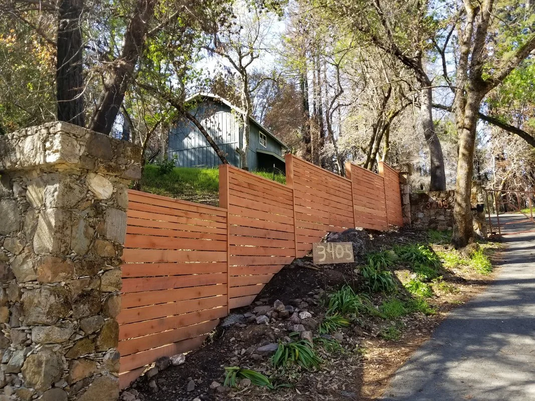 Wooden fence on a hillside with trees and a house in the background, along a paved road.