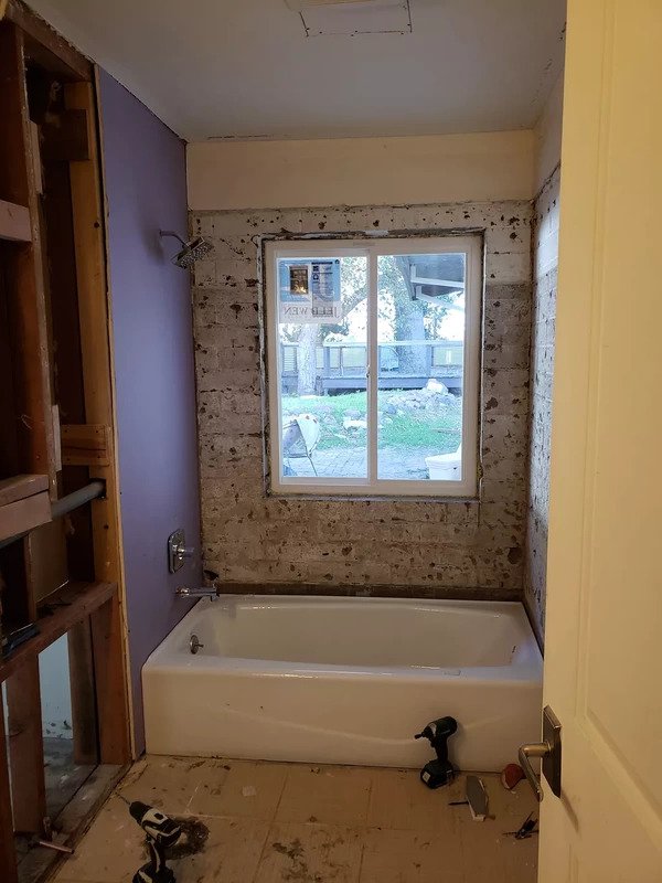 Bathroom under renovation featuring a bathtub beneath a window, with unfinished walls showing exposed brick and insulation, and construction tools on the floor.