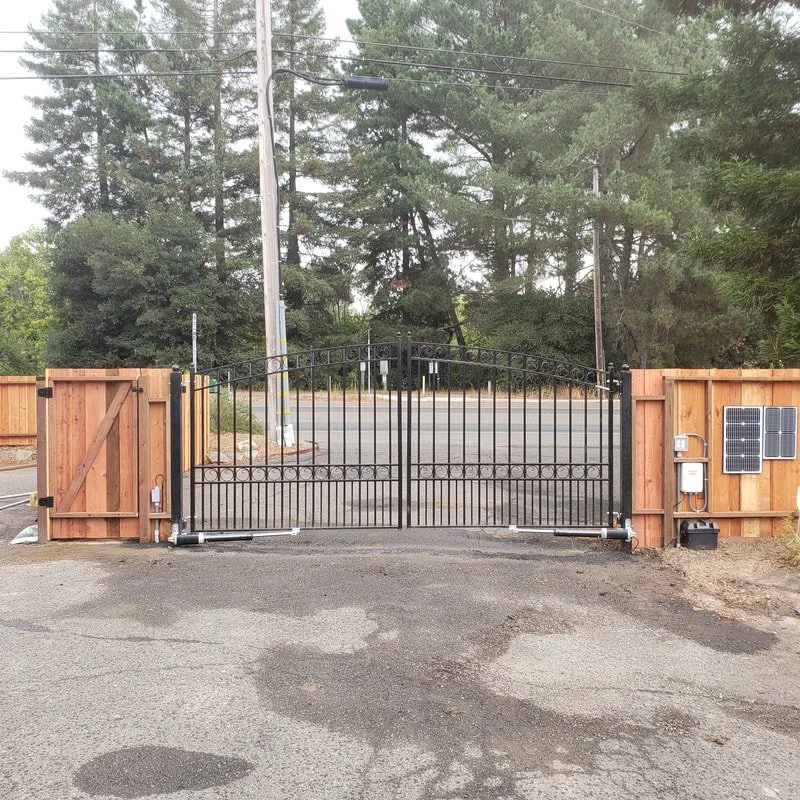 Black metal gate with wooden panels on either side, installed at the entrance of a driveway with a paved surface, surrounded by trees and electrical wires.