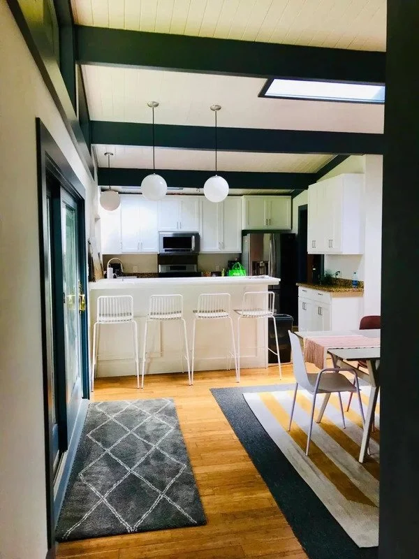 Interior view of a kitchen and dining area with white cabinets, black countertops, and wooden flooring. There are three white barstools at the counter, three pendant lights hanging from the ceiling, and a dining table with chairs on a striped rug. A 