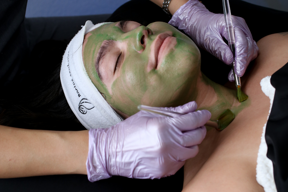Woman receiving a facial treatment with a green mask applied to her face, lying down with her eyes closed, while a skincare professional brushes the mask with a small brush at a skin spa in Henderson NV