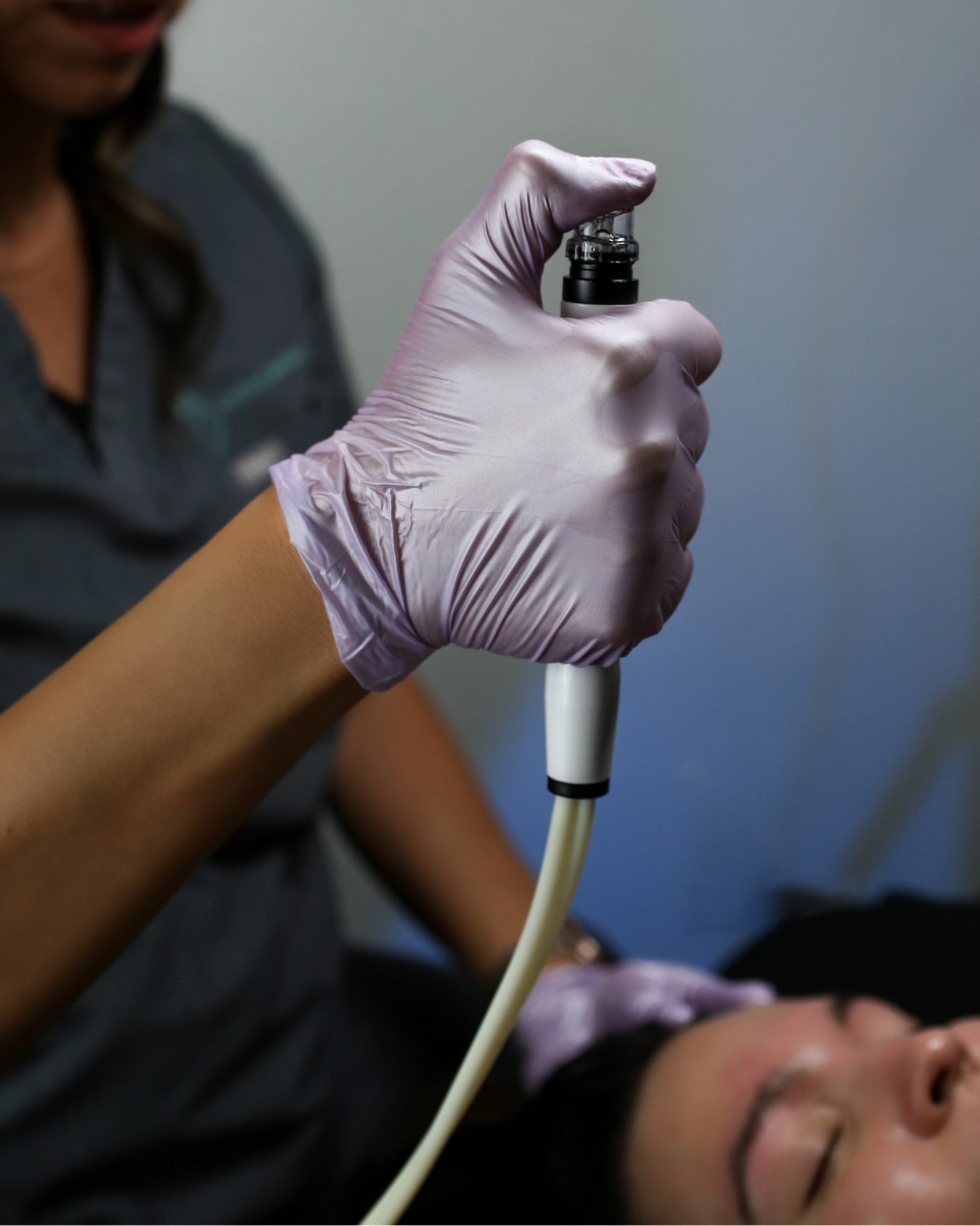 A person wearing a purple glove holding facial suction device called hydrodermabrasion similar to hydrafacial near henderson nevada, with a woman lying down on a spa bed in the background.