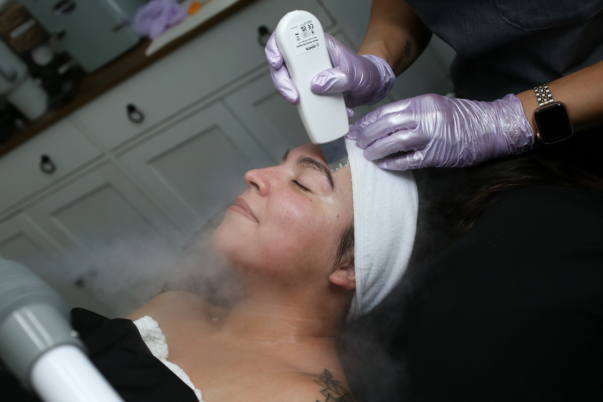 A woman receiving a luxury facial treatment in a Henderson NV spa, lying down with her eyes closed, wearing a headband, while an esthetician applies a cold device to her face.