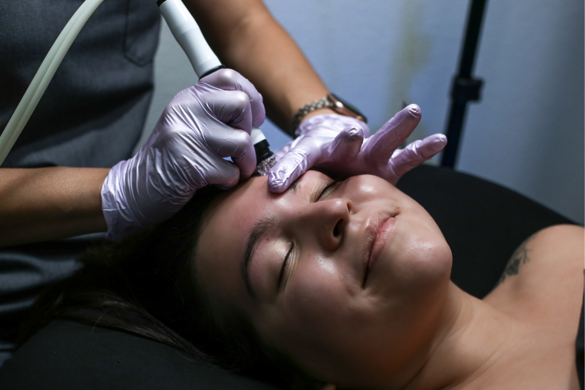 A person receiving a cosmetic facial treatment like a hydrafacial in henderson nv, with a practitioner using a small device on the person's forehead. The practitioner is wearing purple gloves and has a bracelet on one wrist.