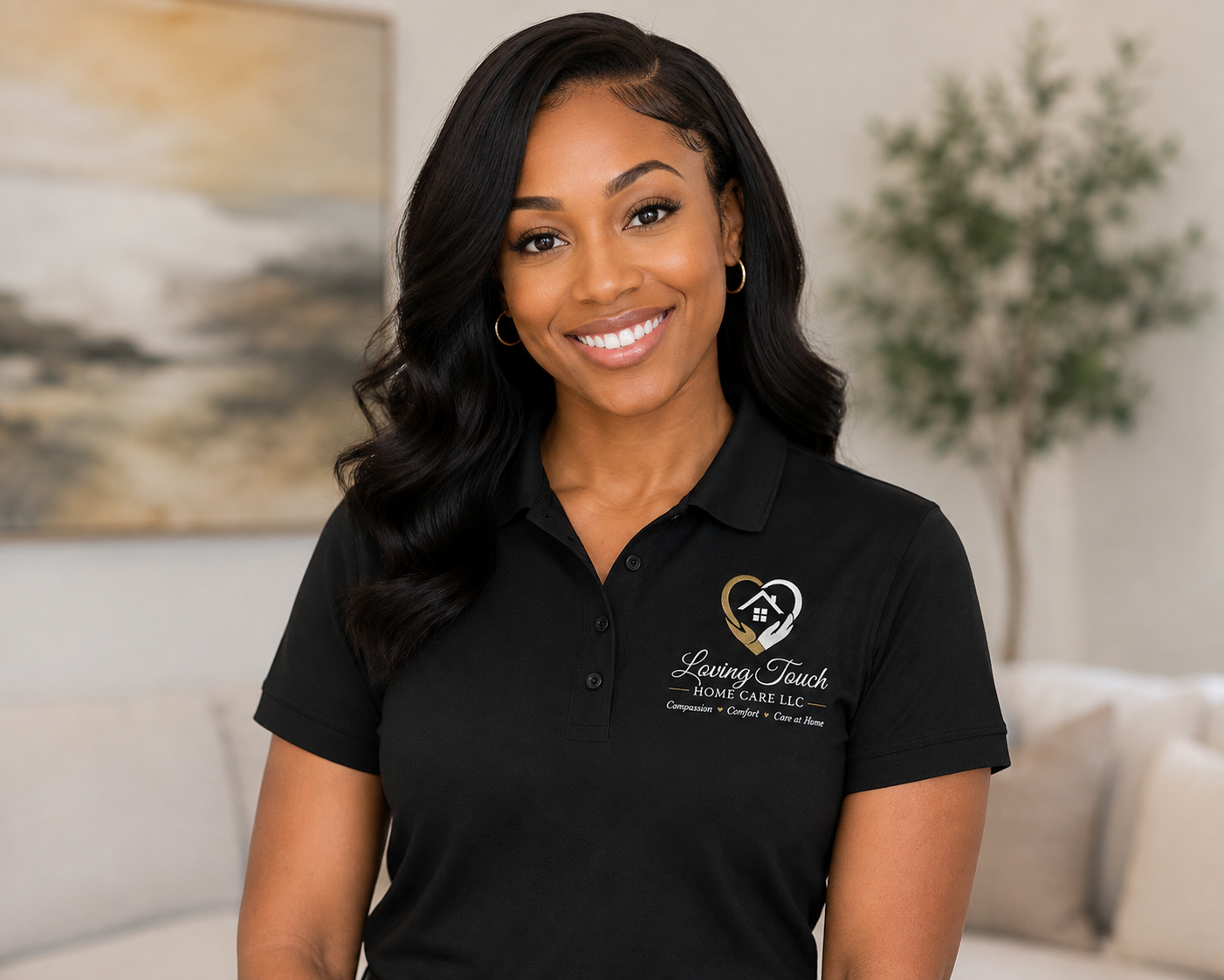 A smiling woman with long black hair wearing a black polo shirt with a logo that says 'Loving Touch Home Care LLC' and a heart with a house inside. The background shows a beige couch, a painting, and a small green plant.