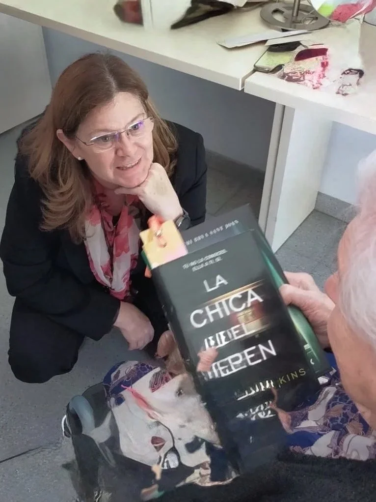 Una mujer está atenta mientras le entrega un libro a una anciana en una residencia como parte de una iniciativa social en apoyo a la soledad que sufren las personas mayores