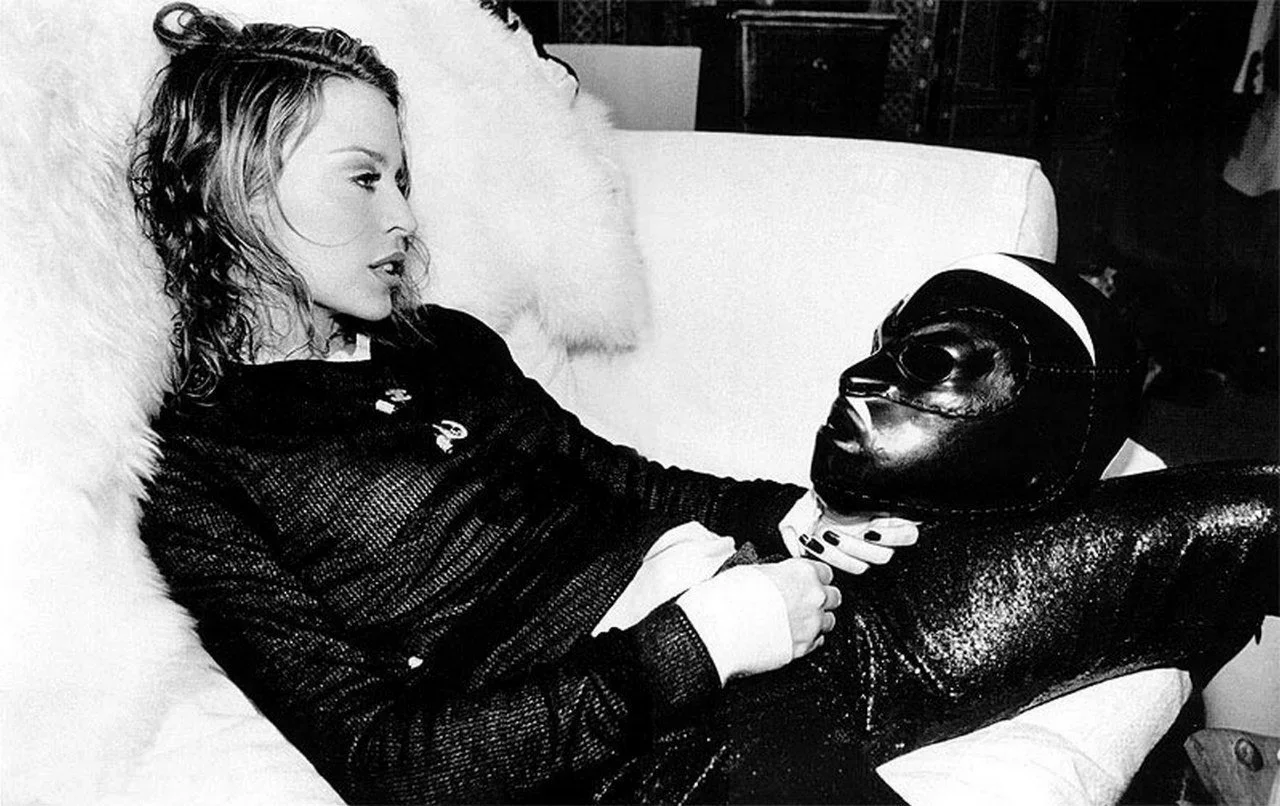 Picture of Kylie Minogue with wavy hair reclining on a bed or couch, wearing a dark outfit, is holding the chin of a leather head sculpture. The scene is in black and white.