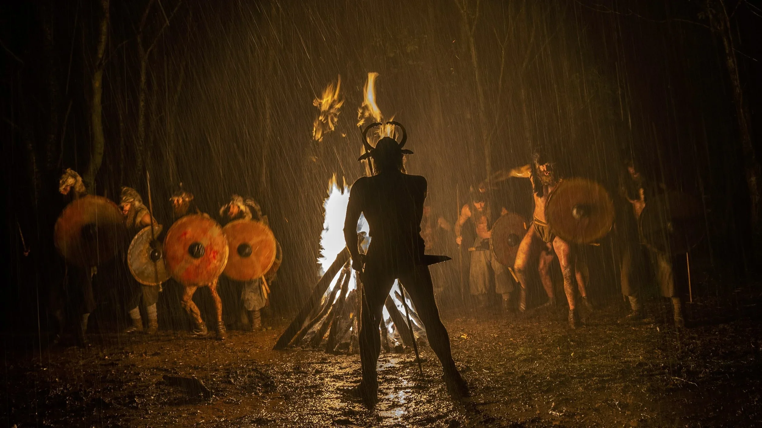 Scene from "The Northman". Silhouette of a person dressed as a Viking with horns on their head standing in front of a bonfire during a rainstorm, surrounded by individuals in Viking costumes holding shields and weapons, in a forest at night.