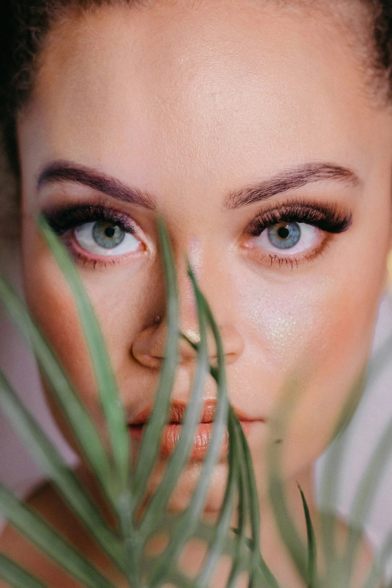 Close-up of a woman's face with light skin and blue eyes, partially obscured by green plant leaves in the foreground.