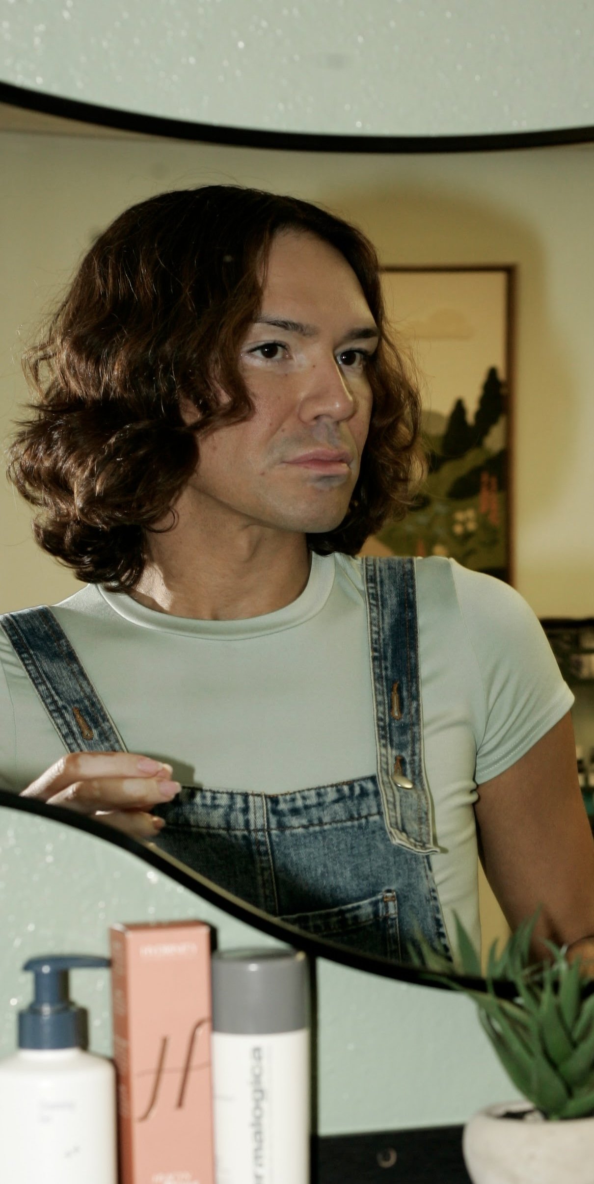 A man with shoulder-length wavy brown hair and light skin looking into a mirror. He is wearing a white t-shirt and denim overalls. The background shows a framed picture on the wall. In the foreground, there are skincare or cosmetic products and a green potted plant.