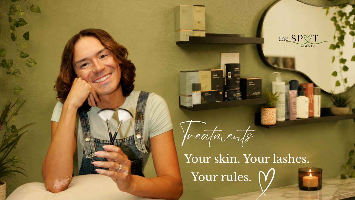 A smiling person with shoulder-length curly hair, wearing a light T-shirt and denim overalls, holding eyelash extension tools in a cozy, green-colored beauty treatment room with shelves of skincare products and a candle.