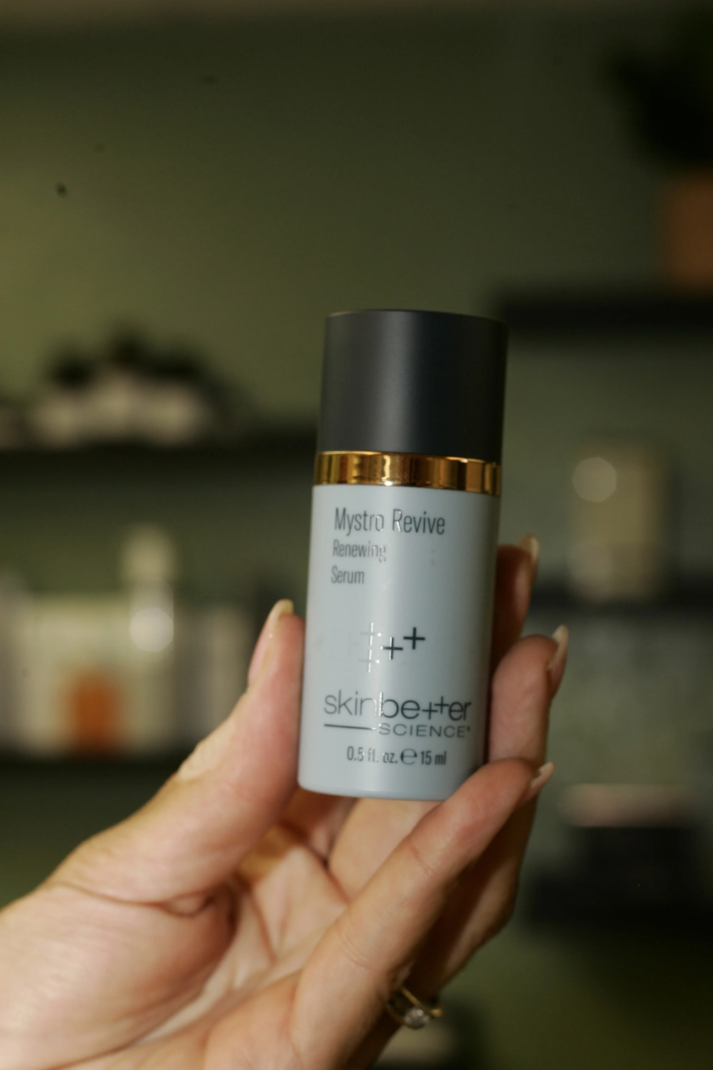 Hand holding a white bottle of SkinBetter Science Mystro Revive serum with a black cap and gold band, blurred background.