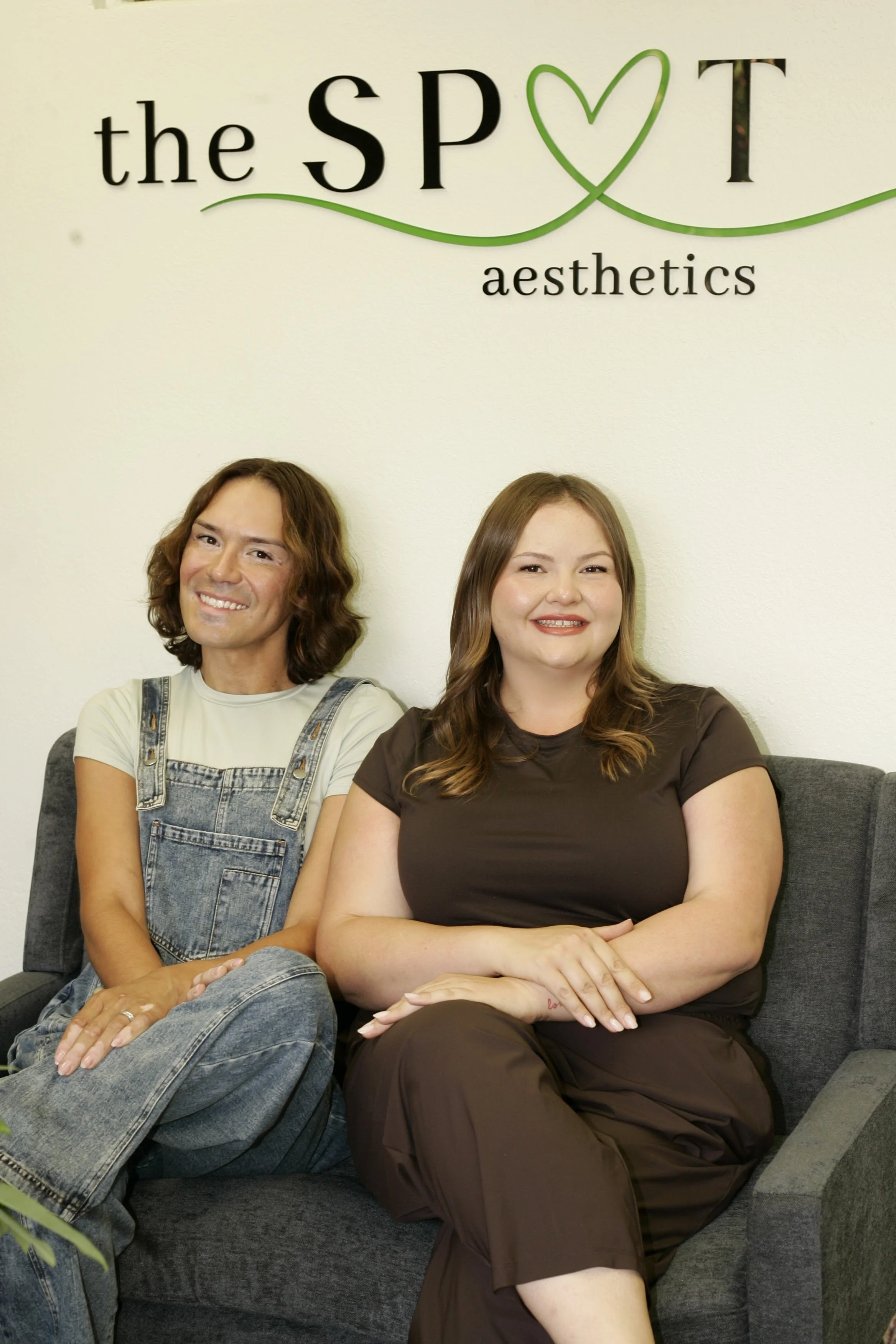 Two women sitting on a gray sofa in front of a white wall with a sign that reads 'the SPOT aesthetics'. The woman on the left is wearing a white t-shirt and denim overalls, with shoulder-length wavy brown hair. The woman on the right is wearing a dark brown t-shirt and matching pants, with long, wavy brown hair.