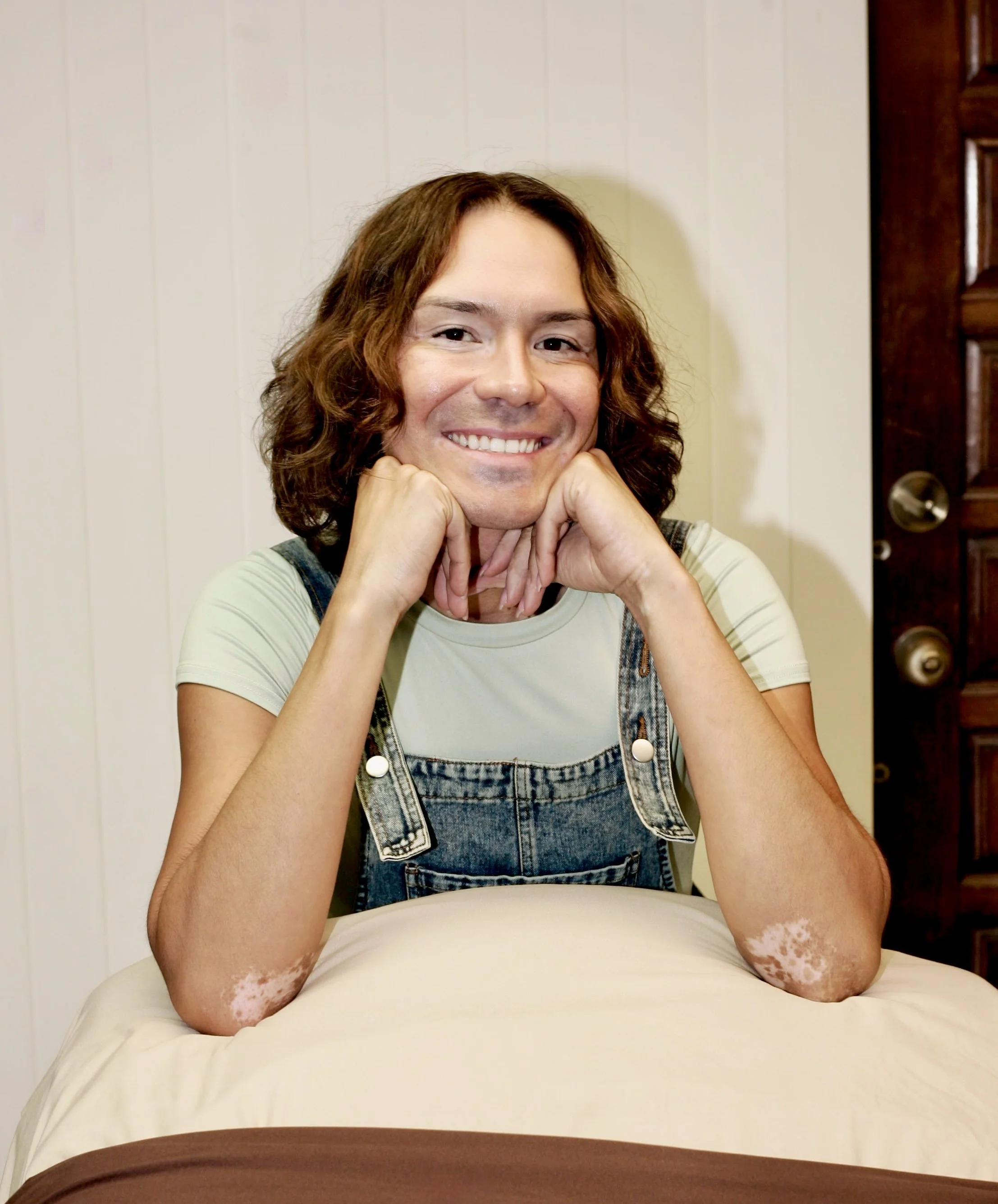 Person with reddish-brown hair smiling with chin resting on hands, wearing a light-colored shirt and denim overalls, sitting on a bed with white sheets, near a wooden door.